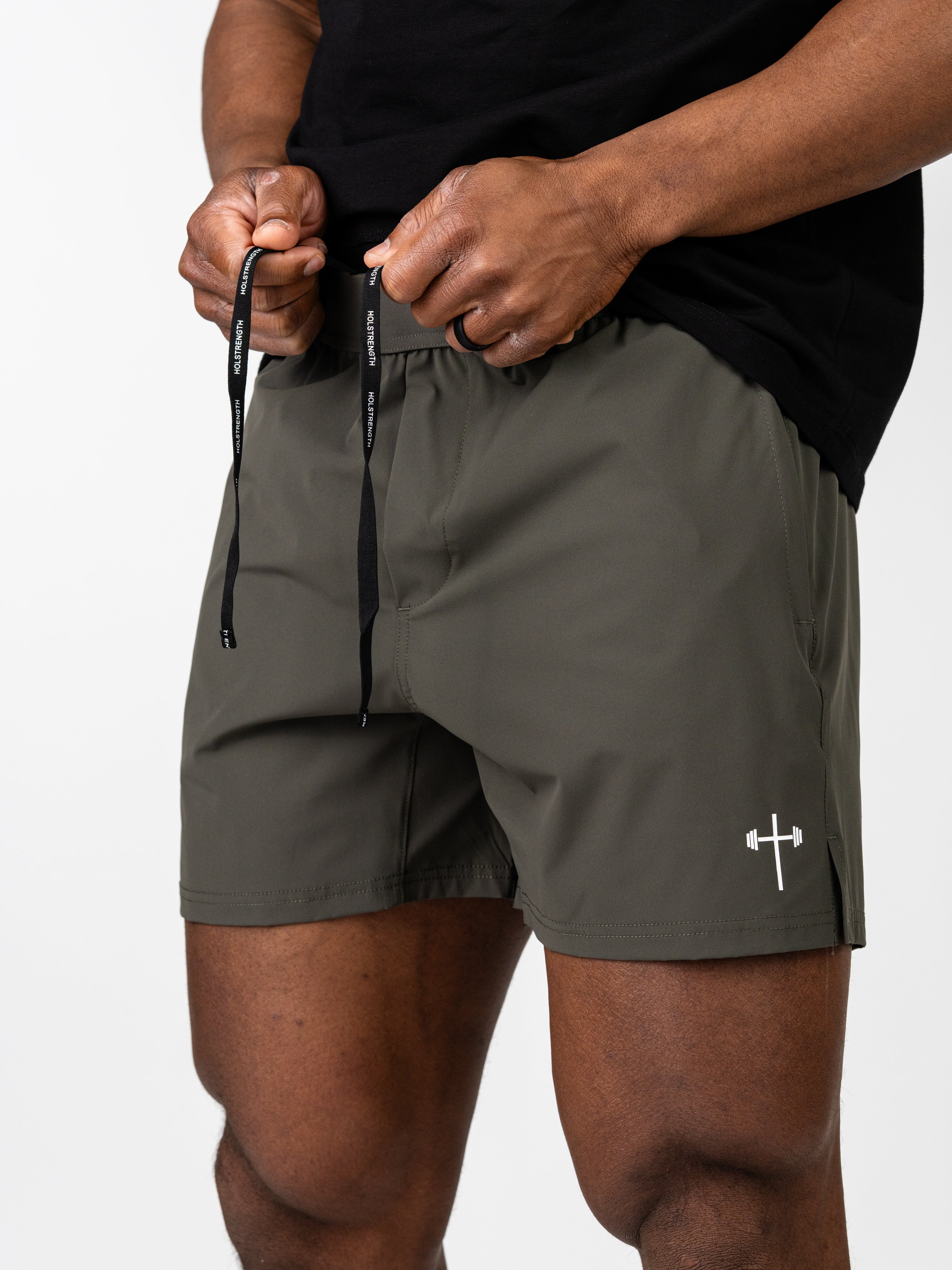 Close-up of a person adjusting the waistband of olive green Essential Training Shorts, featuring a small white barbell cross logo on the left leg, designed for Christian fitness apparel.