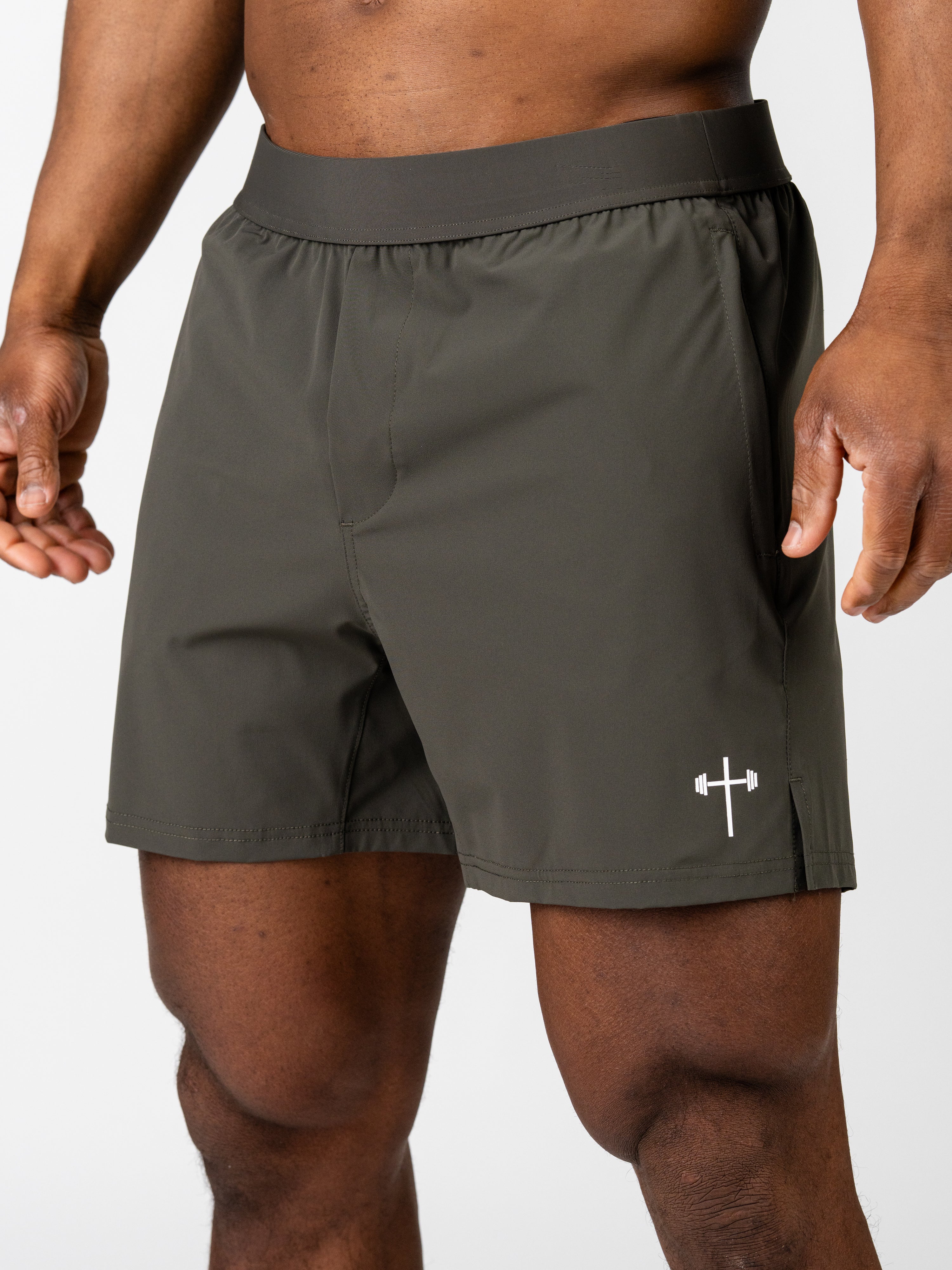 A close-up of a person wearing olive green Essential Training Shorts featuring a lightweight, breathable design and a small white cross with a barbell graphic near the hem.