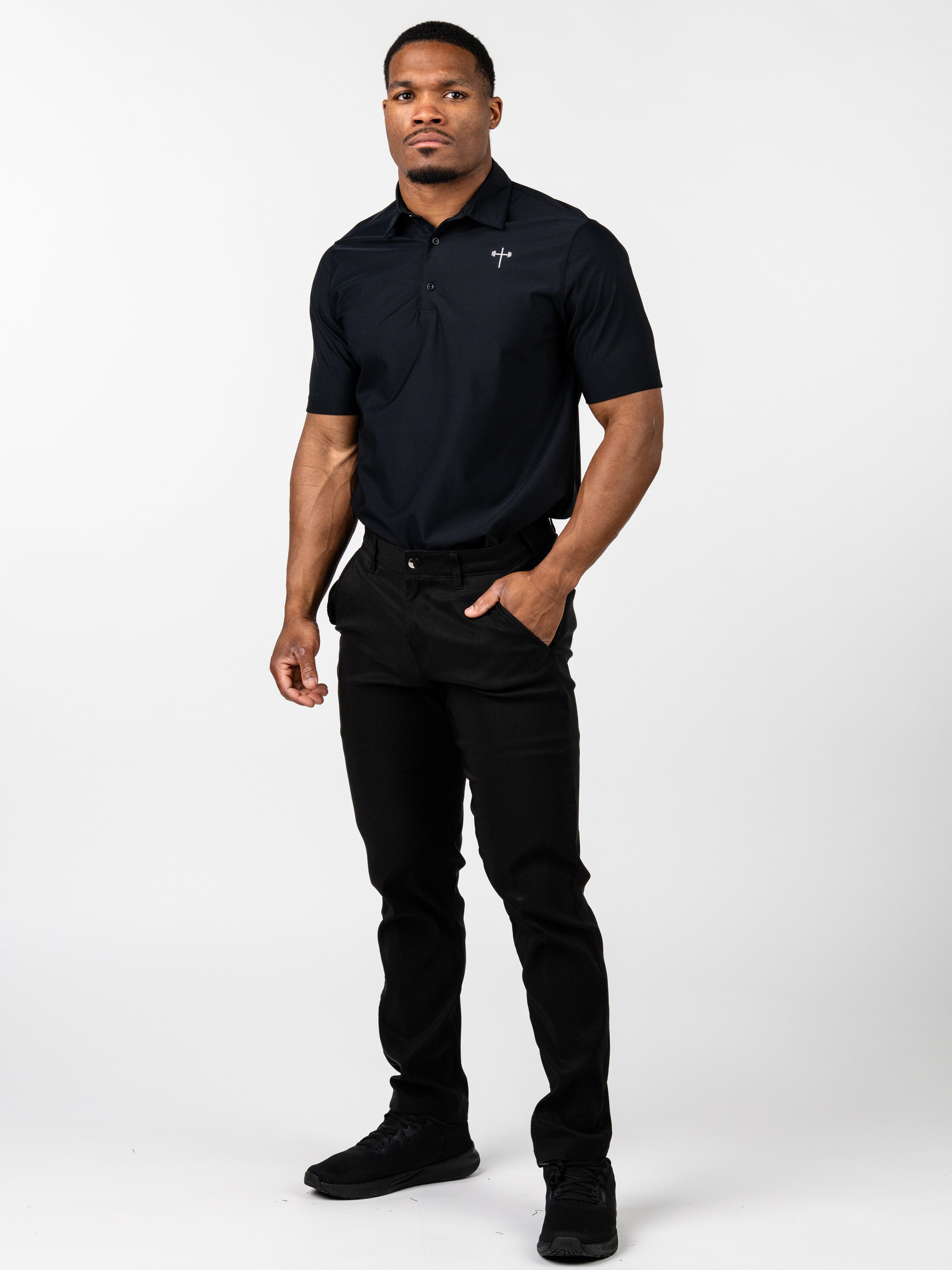 Man wearing a black Core Athletic Polo featuring a subtle Christian cross with barbell design on the chest, paired with black pants and black sneakers, standing against a white background.