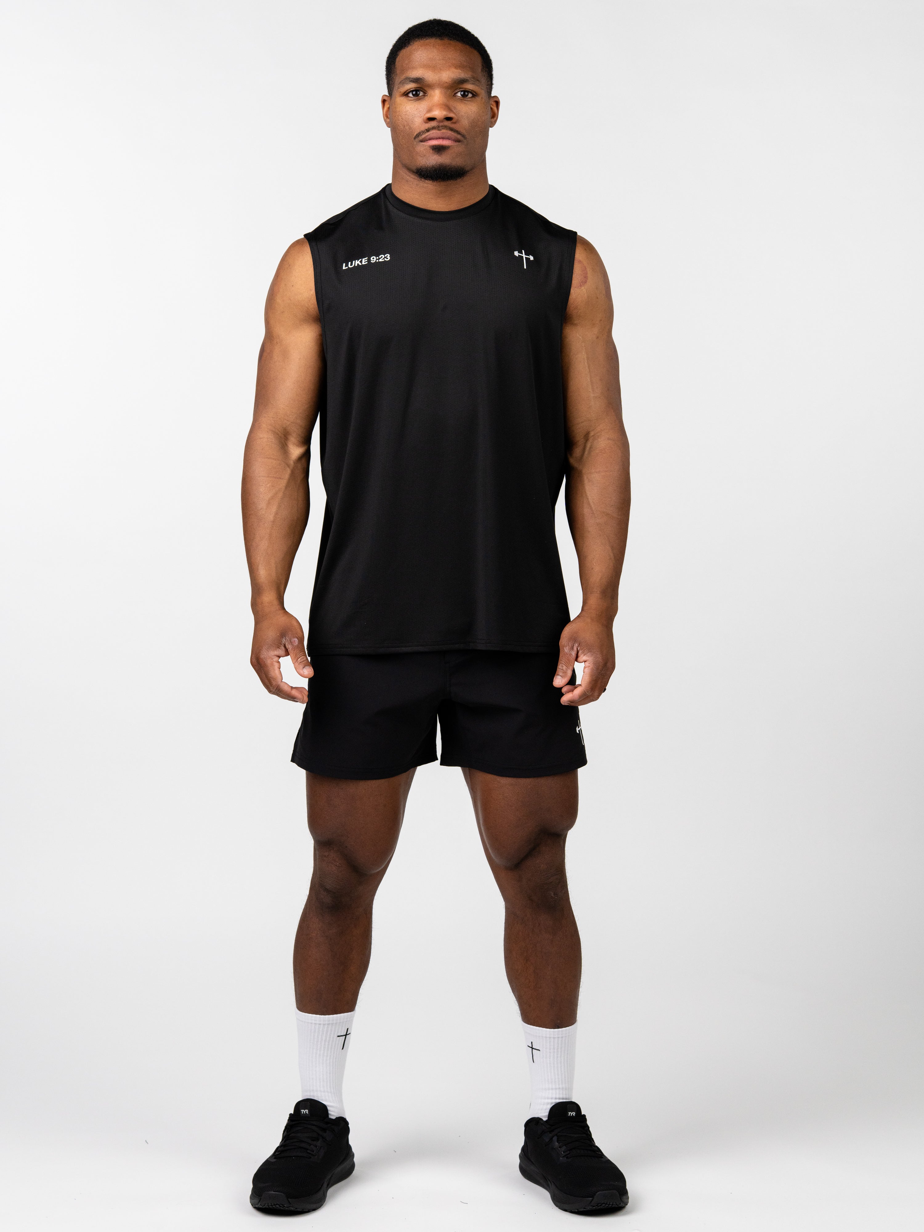 Male model wearing a black Carry Your Cross Premium Oversized Tank with subtle cross and scripture details, paired with black athletic shorts, socks, and shoes.