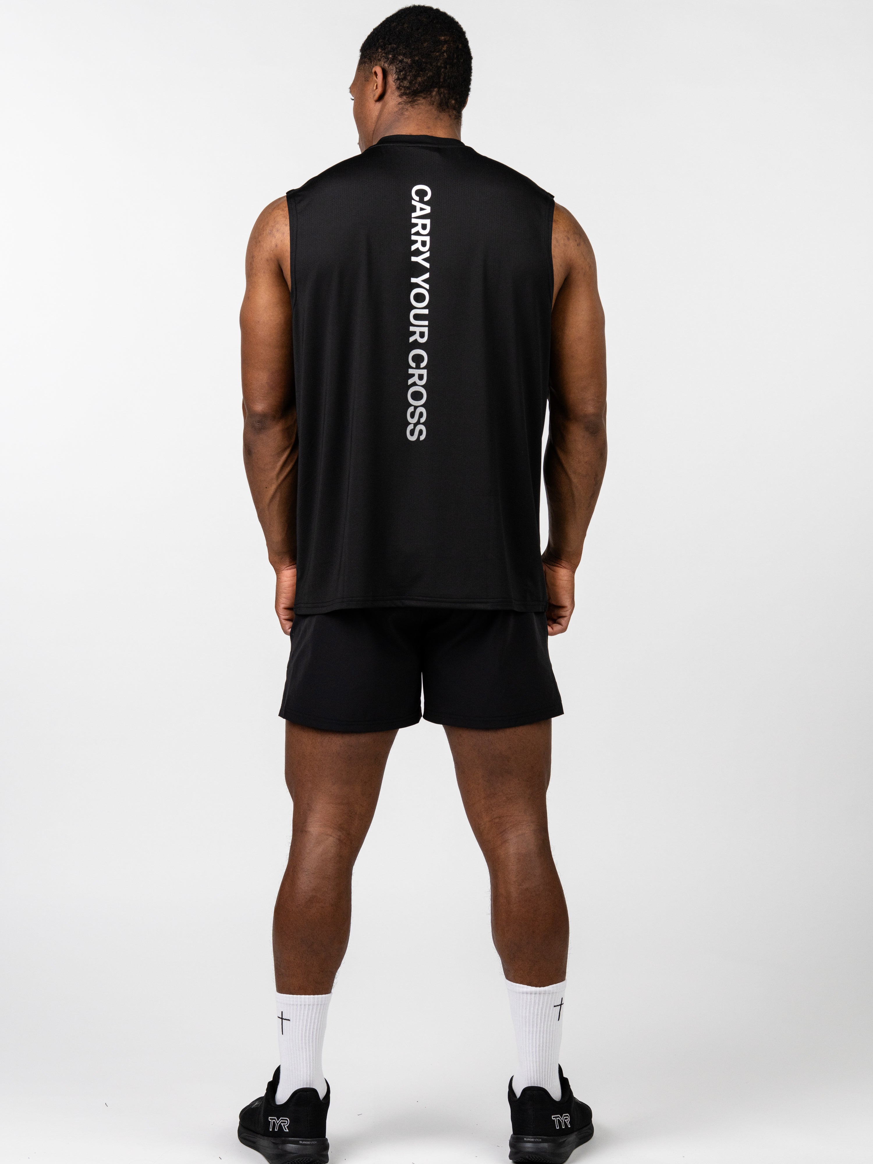 Man wearing the Carry Your Cross Premium Oversized Tank in black, showing the back with vertical "CARRY YOUR CROSS" text, paired with black shorts and white socks with cross detail, representing Christian fitness apparel.