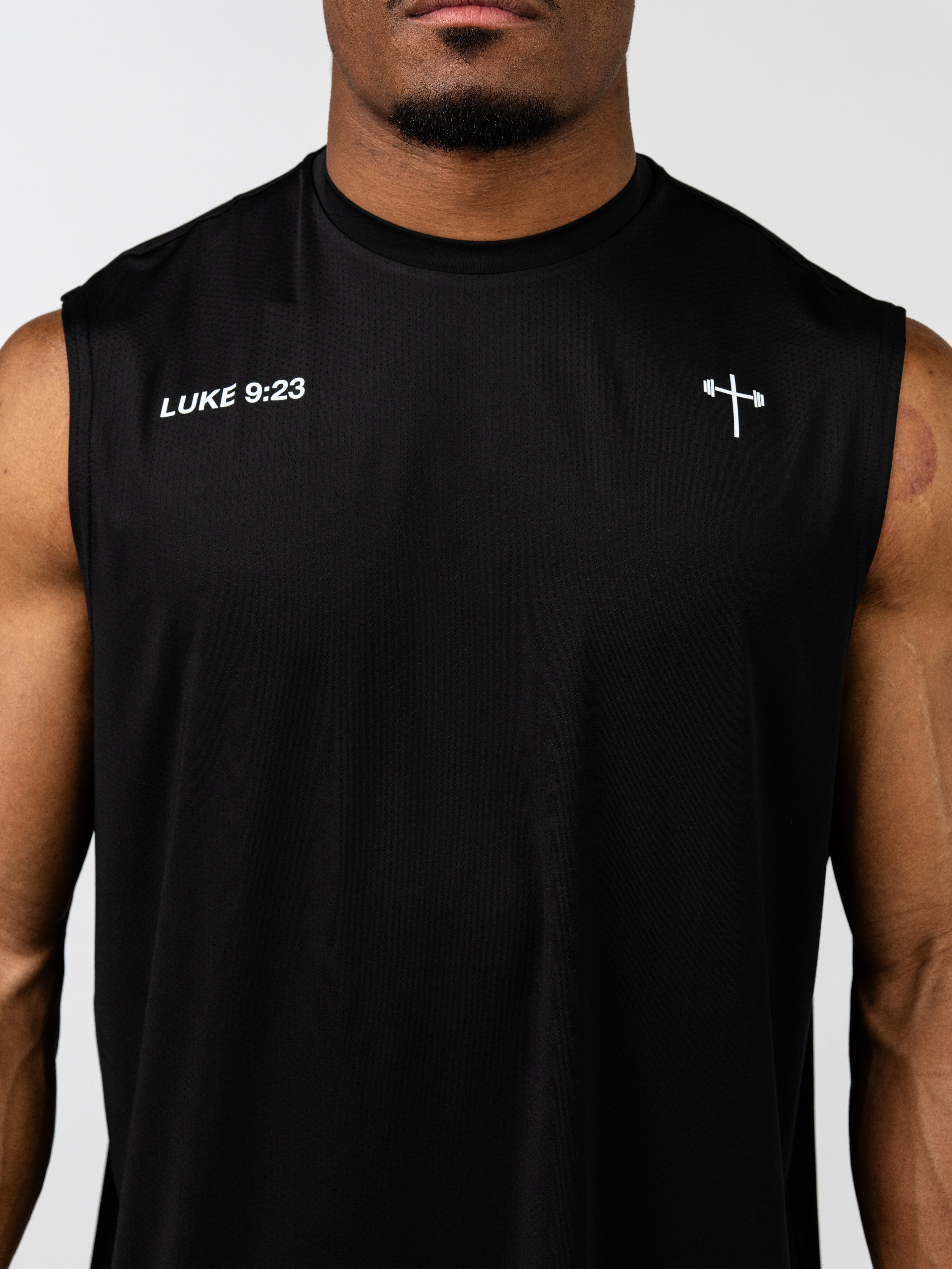 Close-up of a man wearing a black Carry Your Cross Premium Oversized Tank with a small white cross and barbell symbol on the chest and the text LUKE 9:23 on the upper left side, representing Christian gym clothing and faith based fitness apparel.