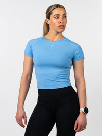 Woman wearing a light blue Women's Combat Tee with a small white cross and barbell design on the chest, paired with black workout leggings and a white fitness watch.