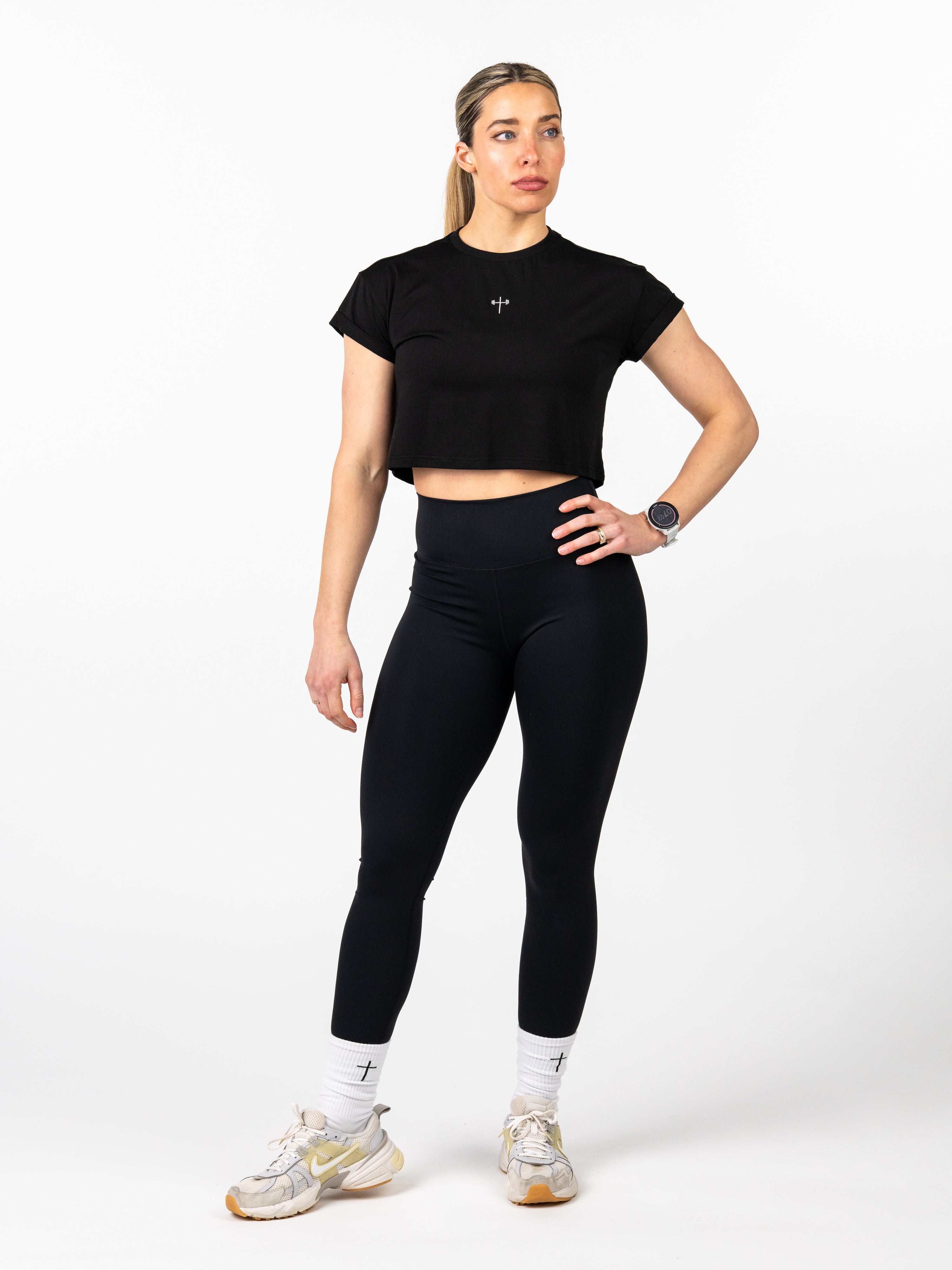 Woman modeling the Cross Essential Crop Tee, a black cropped performance tee with a small subtle cross design at the center chest, styled with black leggings and white socks featuring a cross.