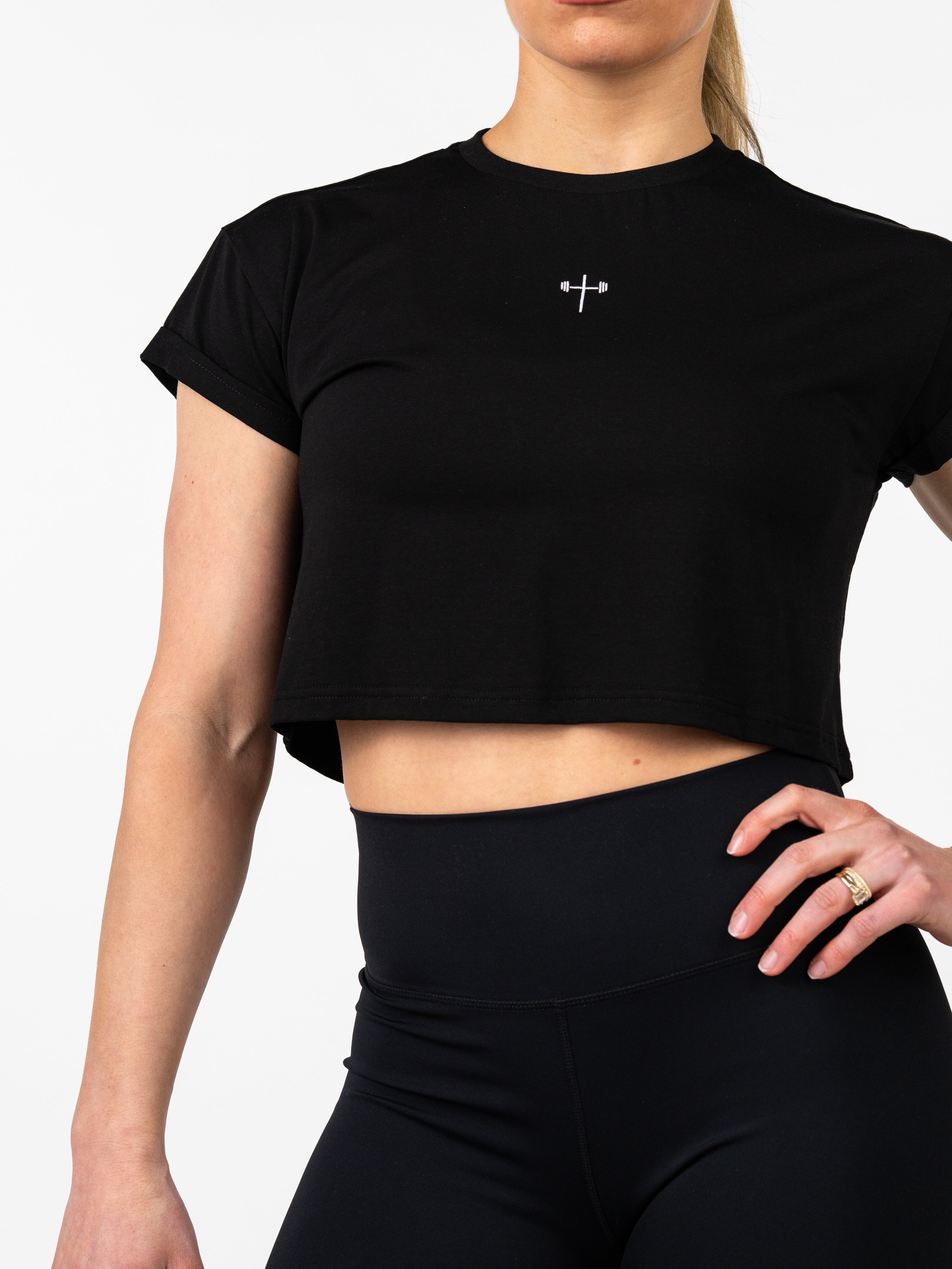 Woman wearing a black Cross Essential Crop Tee featuring a small white cross with a barbell design on the chest, paired with black high-waisted leggings for fitness and casual wear.