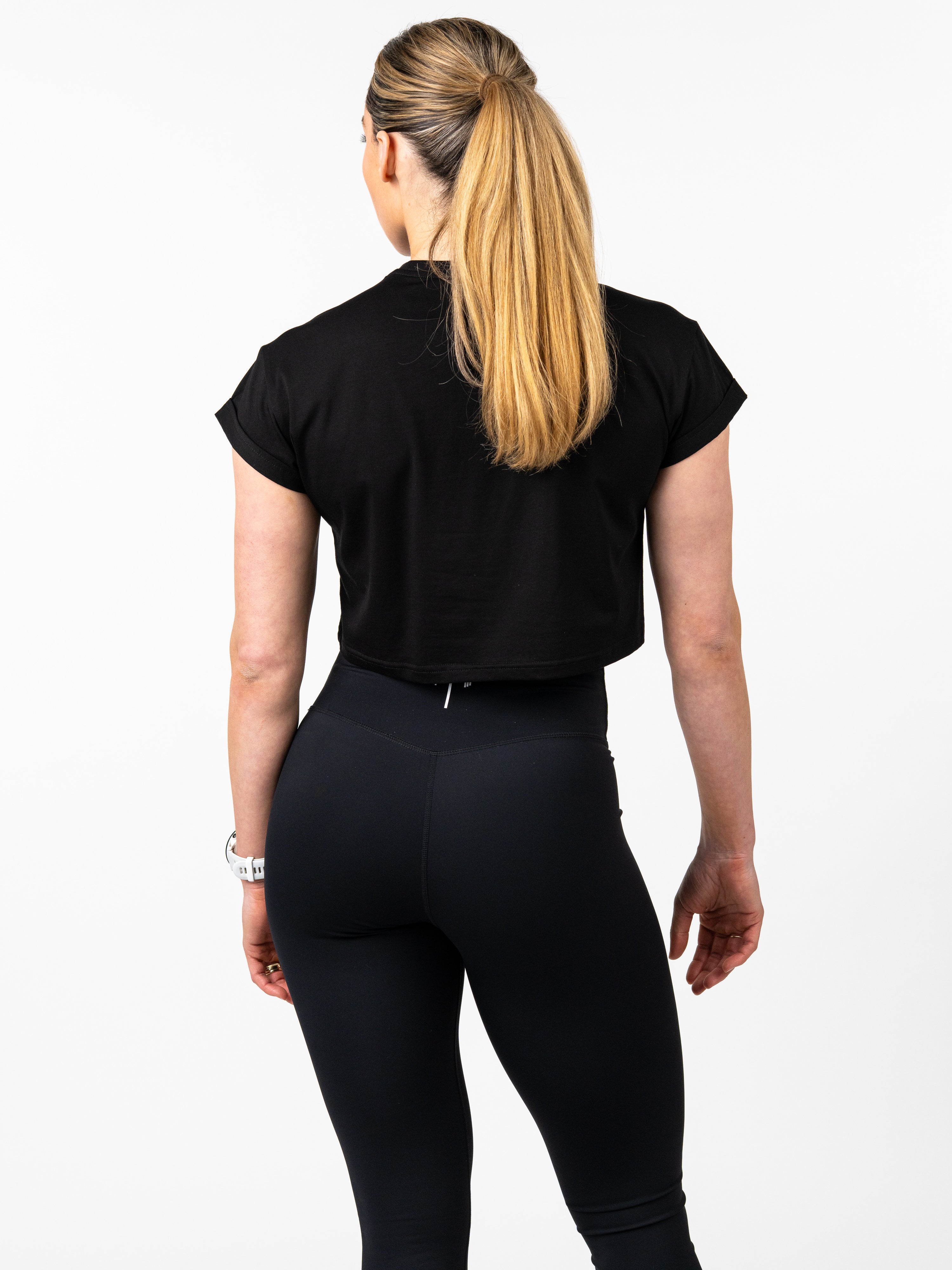 Woman wearing a black Cross Essential Crop Tee and black high-waisted leggings, shown from the back, highlighting the crop tee’s minimal design and comfortable fit for Christian fitness apparel.