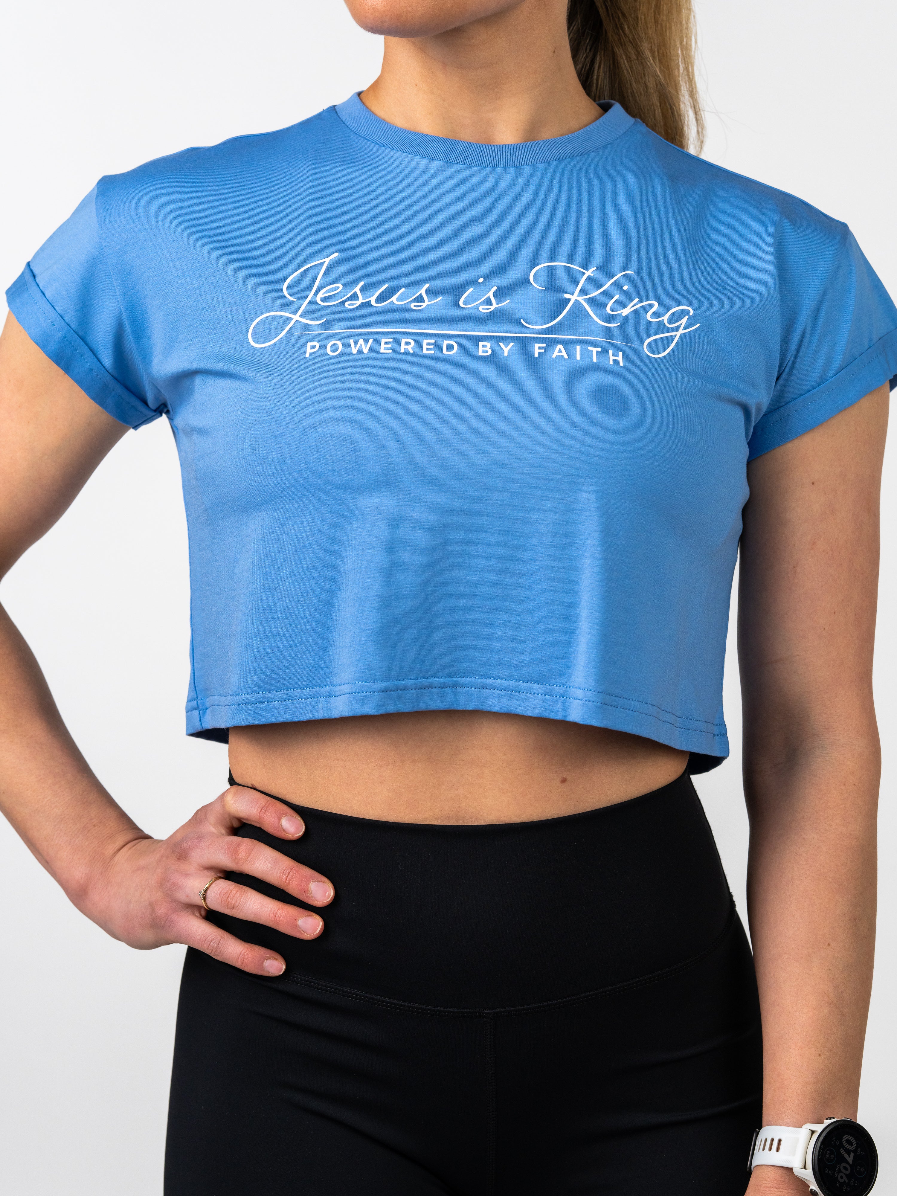 Woman wearing a blue Jesus is King Essential Crop Tee with white script text, paired with black leggings, styled for Christian fitness apparel and faith-based activewear.