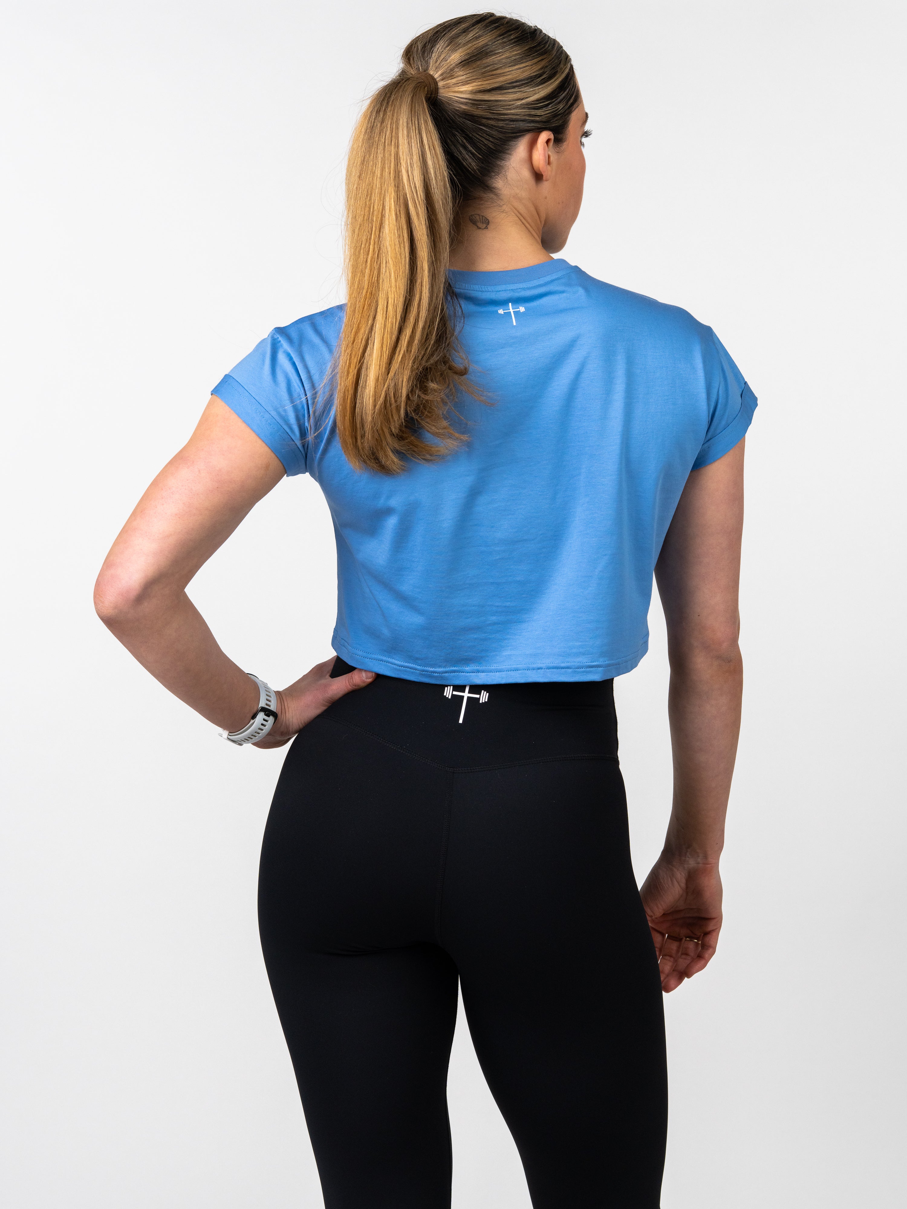 Woman wearing a blue Jesus is King Essential Crop Tee paired with black leggings, showcasing the back design of the Christian fitness apparel in a studio setting.
