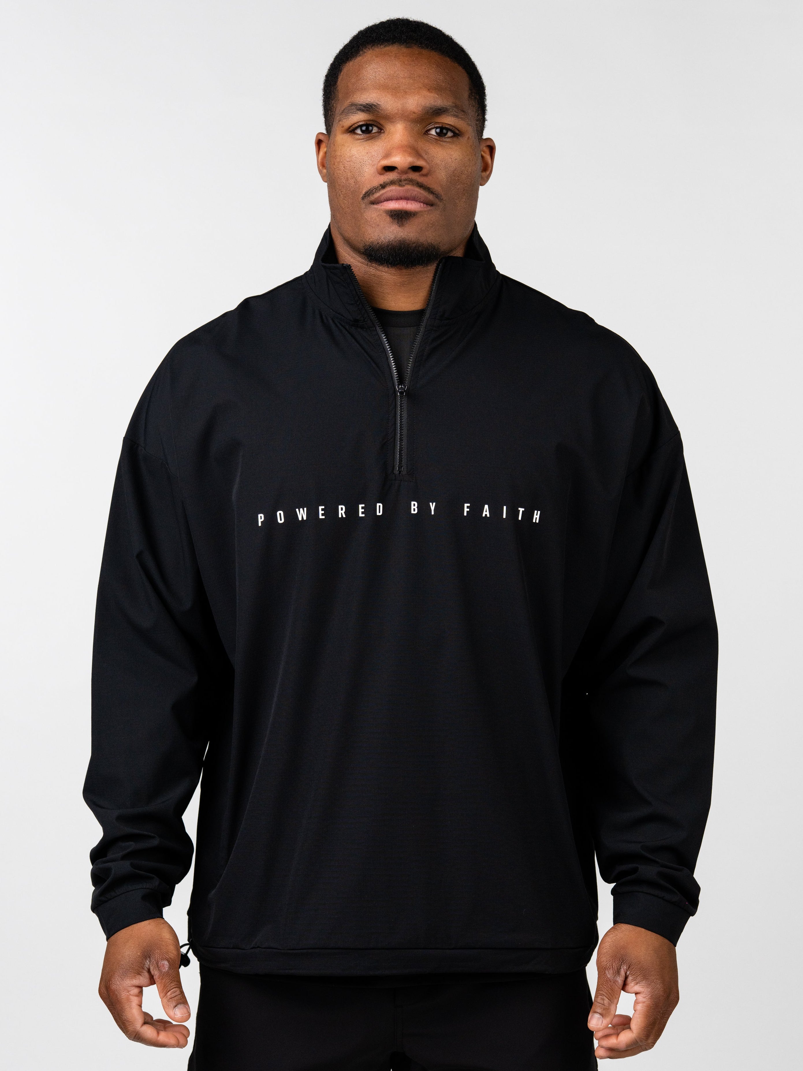 Man wearing a black Powered By Faith Pullover Jacket with a front zipper and white lettering across the chest, styled for Christian fitness apparel.