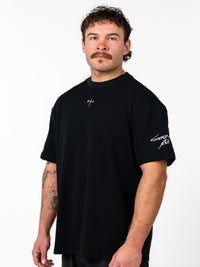 Man wearing a black Sacred Heart Premium Oversized Tee with a small cross and barbell design on the chest and "Sacred Jesus" text on the sleeve, showcasing faith based fitness apparel.