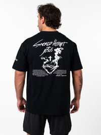 Man wearing black Sacred Heart Premium Oversized Tee with white graphic of a crowned heart, cross, barbell design, and biblical text on the back, styled as Christian fitness apparel.