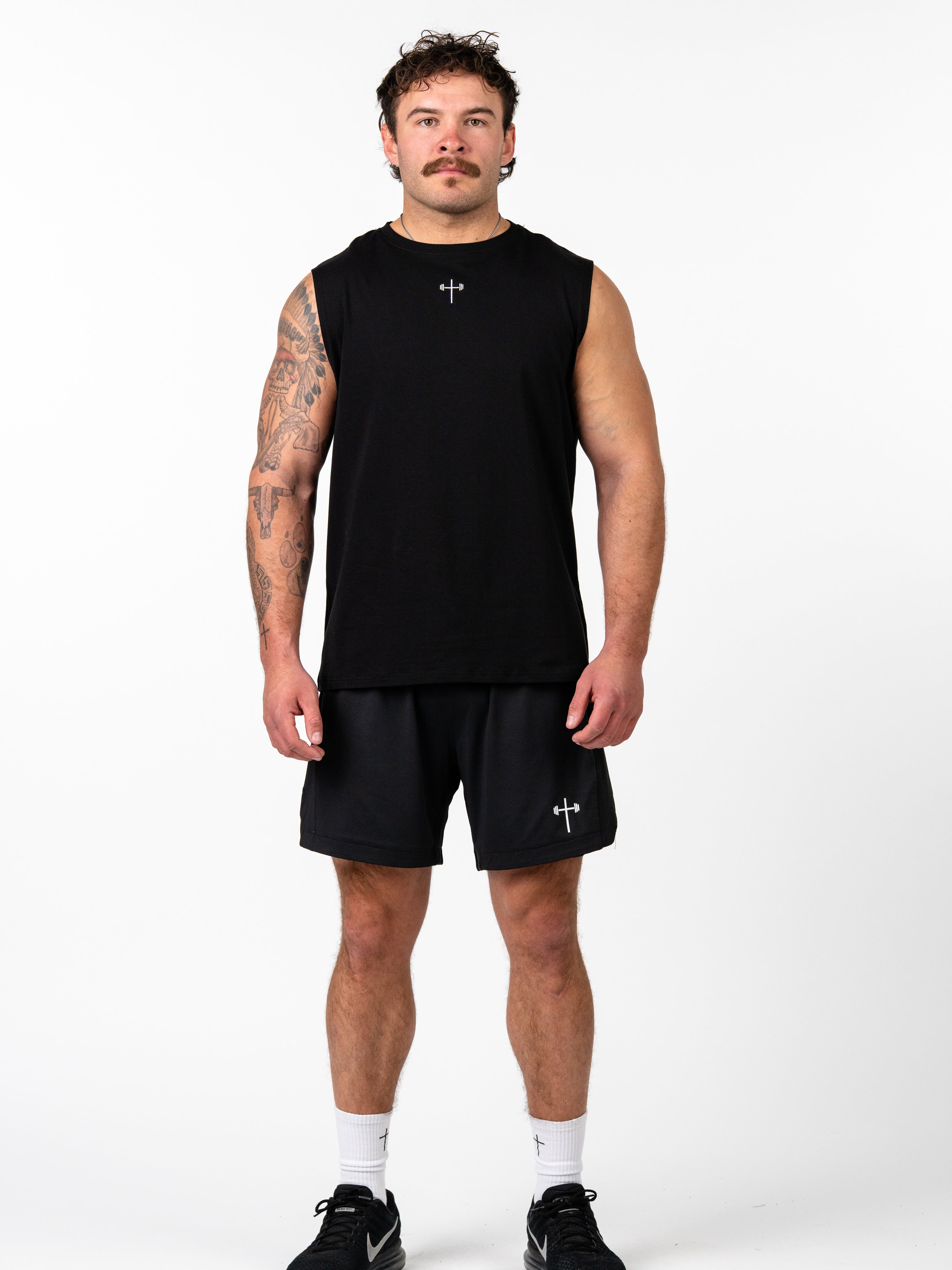 Man wearing Sacred Heart Premium Sleeveless Tank in black with a small Christian cross and barbell design, paired with matching black shorts and athletic shoes.