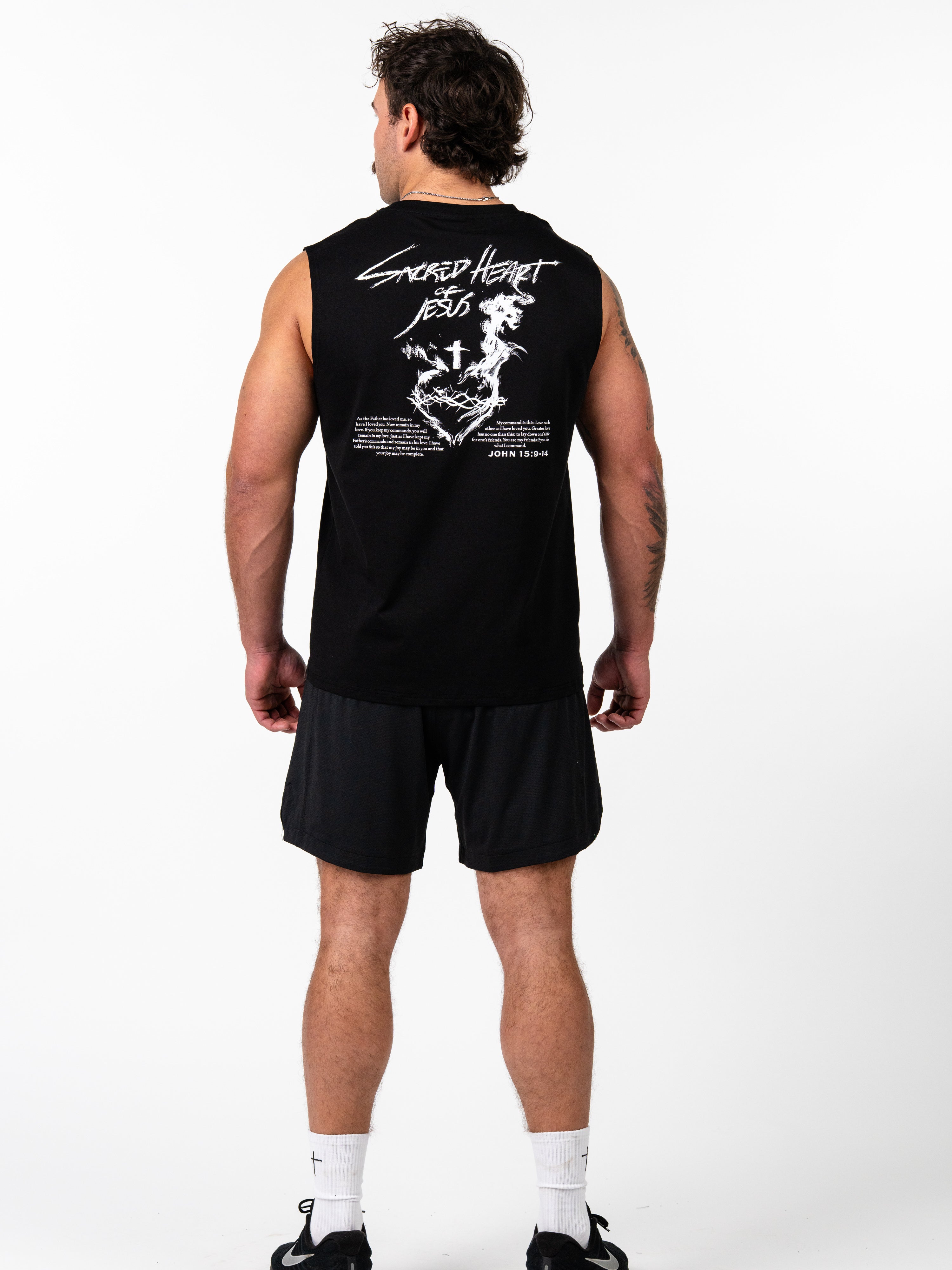 Man wearing black Sacred Heart Premium Sleeveless Tank with white graphic design featuring a sacred heart, cross, and scripture text on the back, paired with black shorts and athletic shoes.