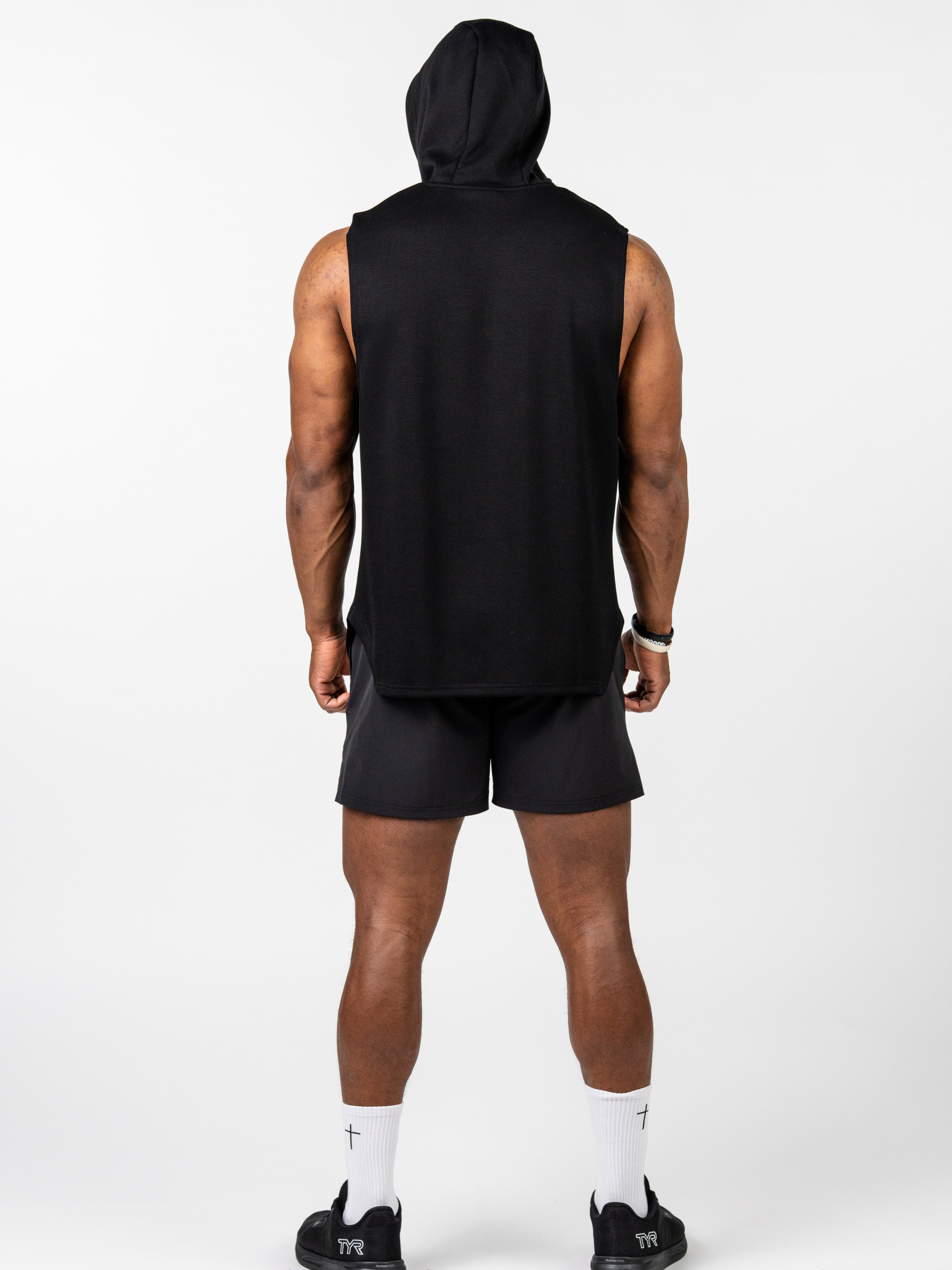 Rear view of a man wearing a black Essential Sleeveless Hoodie with a hood up, paired with black shorts, white socks featuring a cross design, and black athletic shoes.