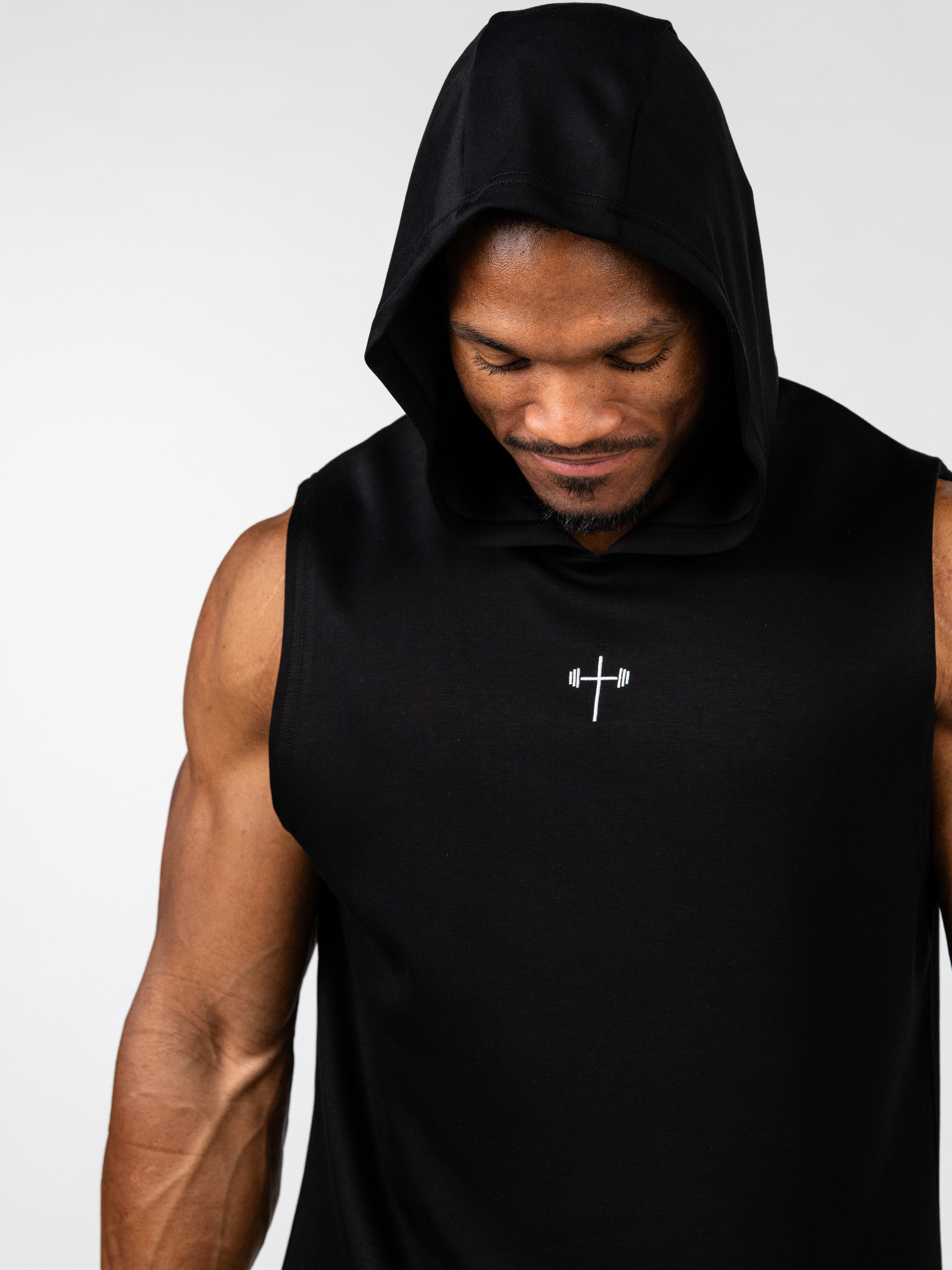 Man wearing a black Essential Sleeveless Hoodie with hood up, featuring a small white cross with barbells graphic on the chest, showcasing Christian fitness apparel.