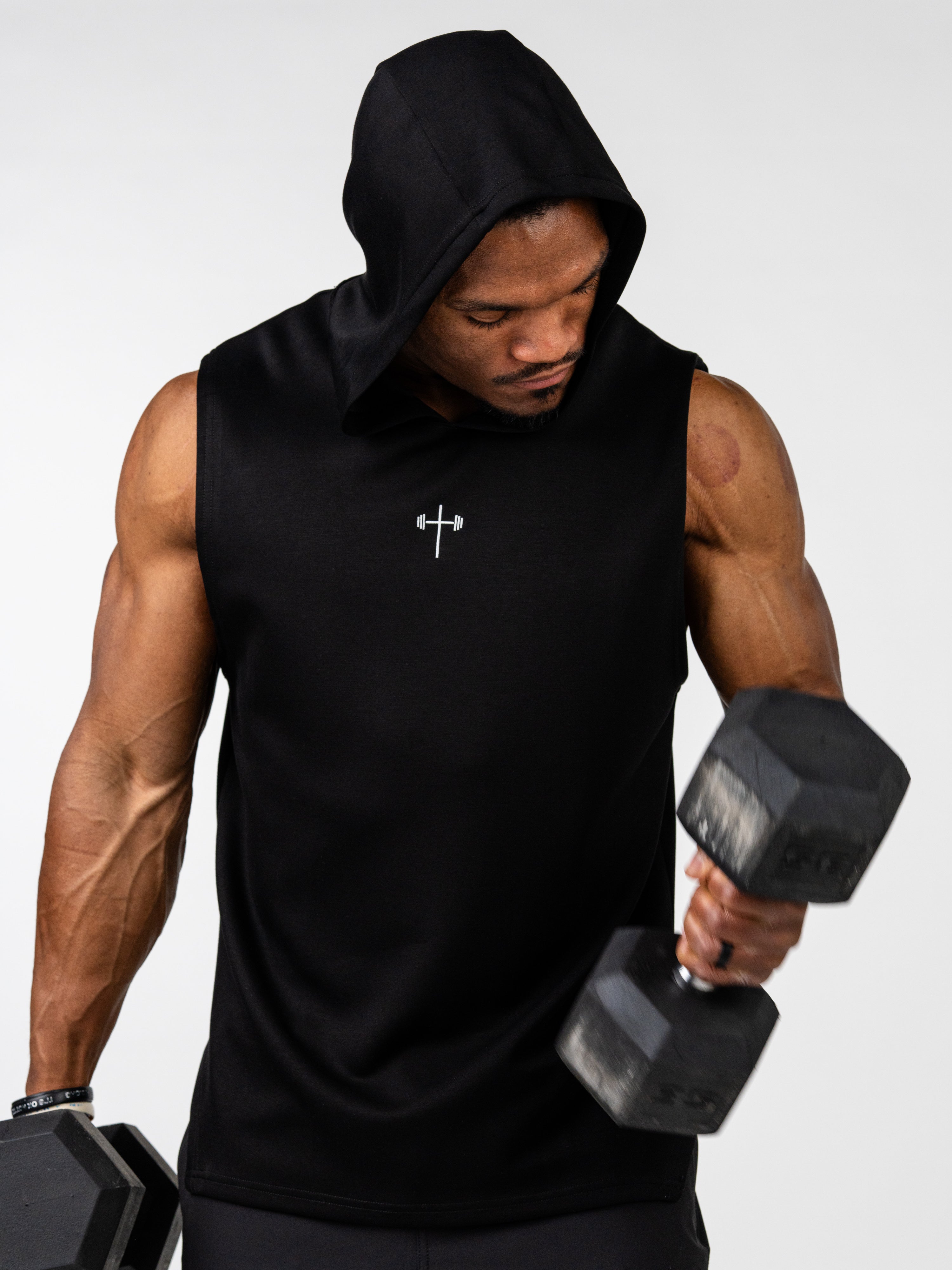 Man wearing a black Essential Sleeveless Hoodie with a subtle cross and barbell design lifting dumbbells, showcasing muscular arms and fitness-focused Christian gym clothing.