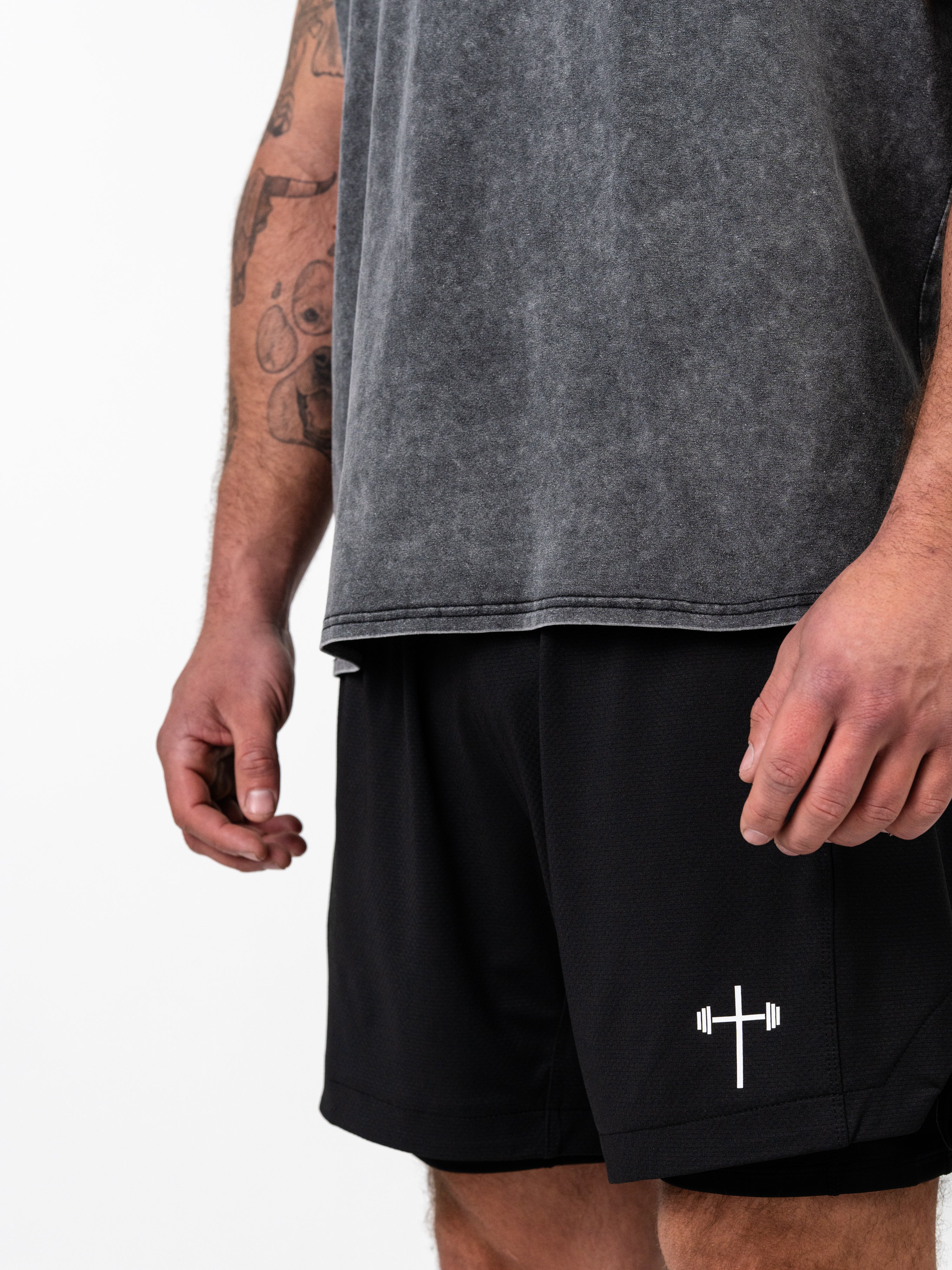 Close-up of a man wearing God is Love Vintage Premium Tank in faded gray paired with black athletic shorts featuring a white cross and barbell symbol, representing Christian fitness apparel.