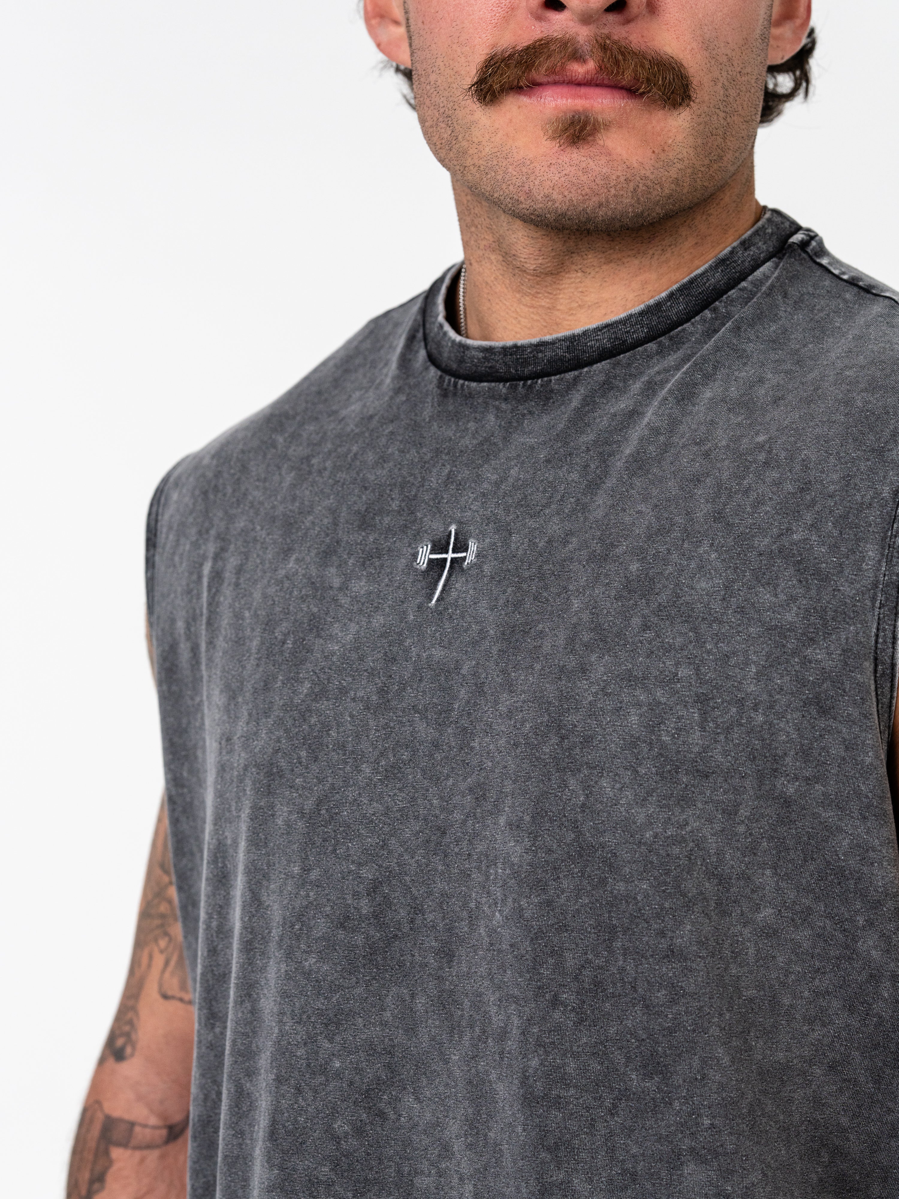 Man wearing a faded black God is Love Vintage Premium Tank featuring a small embroidered cross with a barbell design on the chest, representing Christian fitness apparel.