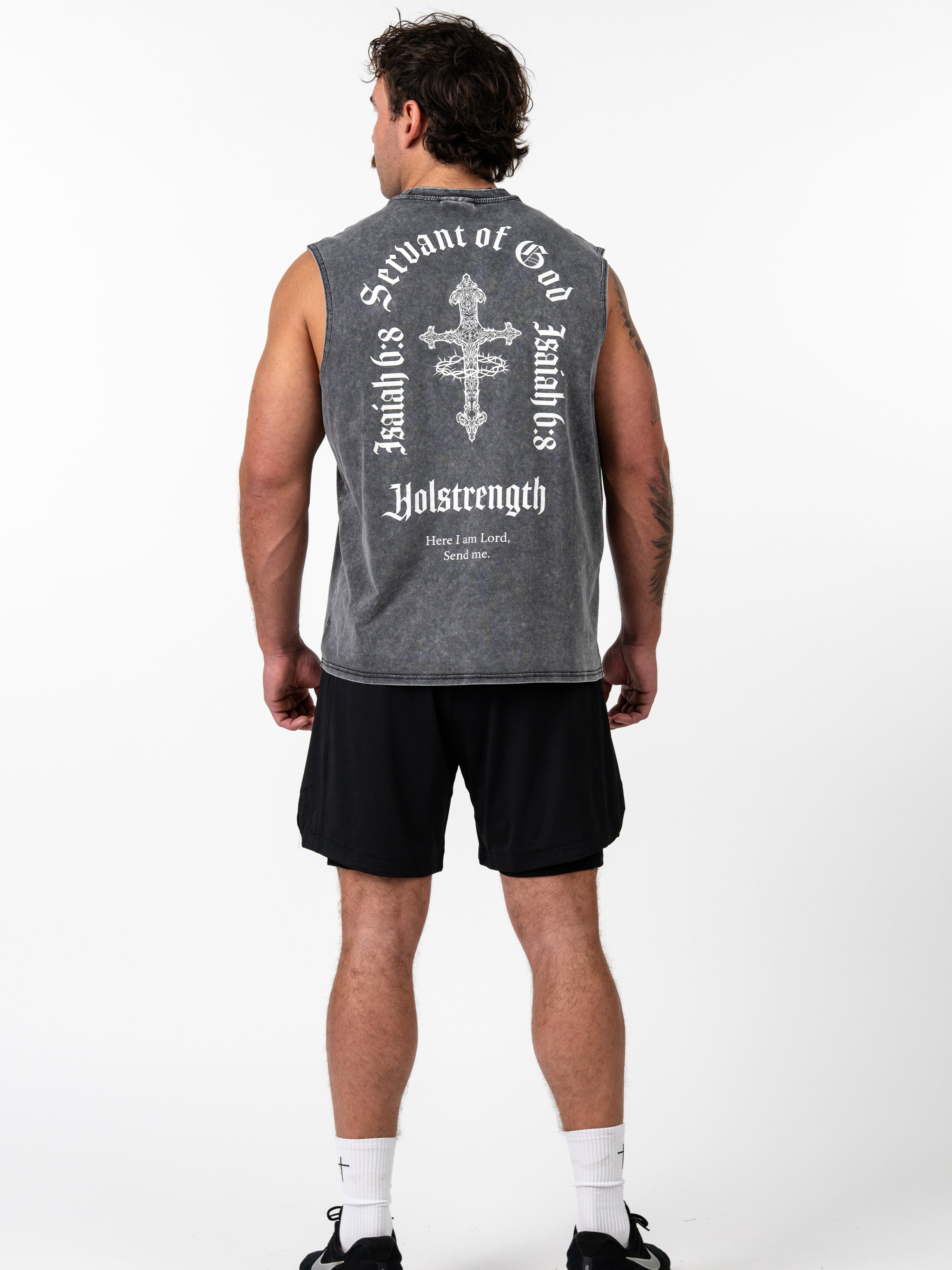 Man wearing a dark gray sleeveless Servant of God Vintage Premium Tank with a large white cross and Isaiah 6:8 scripture text on the back, paired with black shorts and athletic shoes.