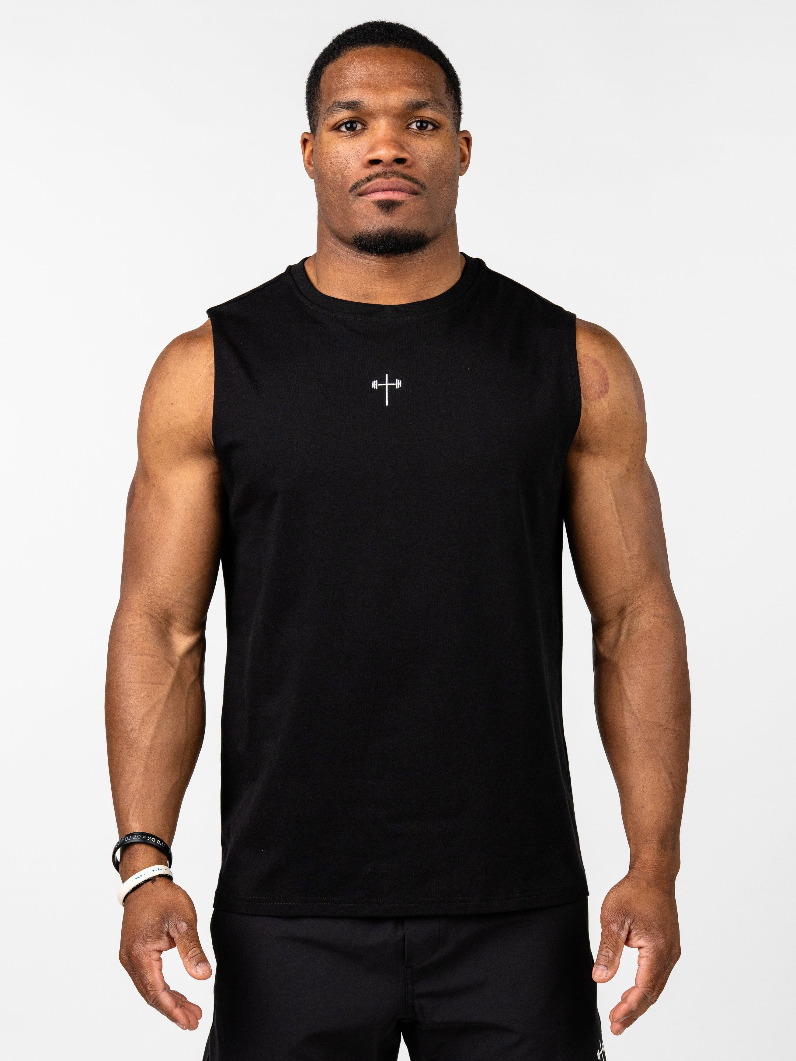 Athletic man wearing a black Vital Cross Sleeveless Tank featuring a small cross with a barbell design in the center, styled for high-output training and comfort.