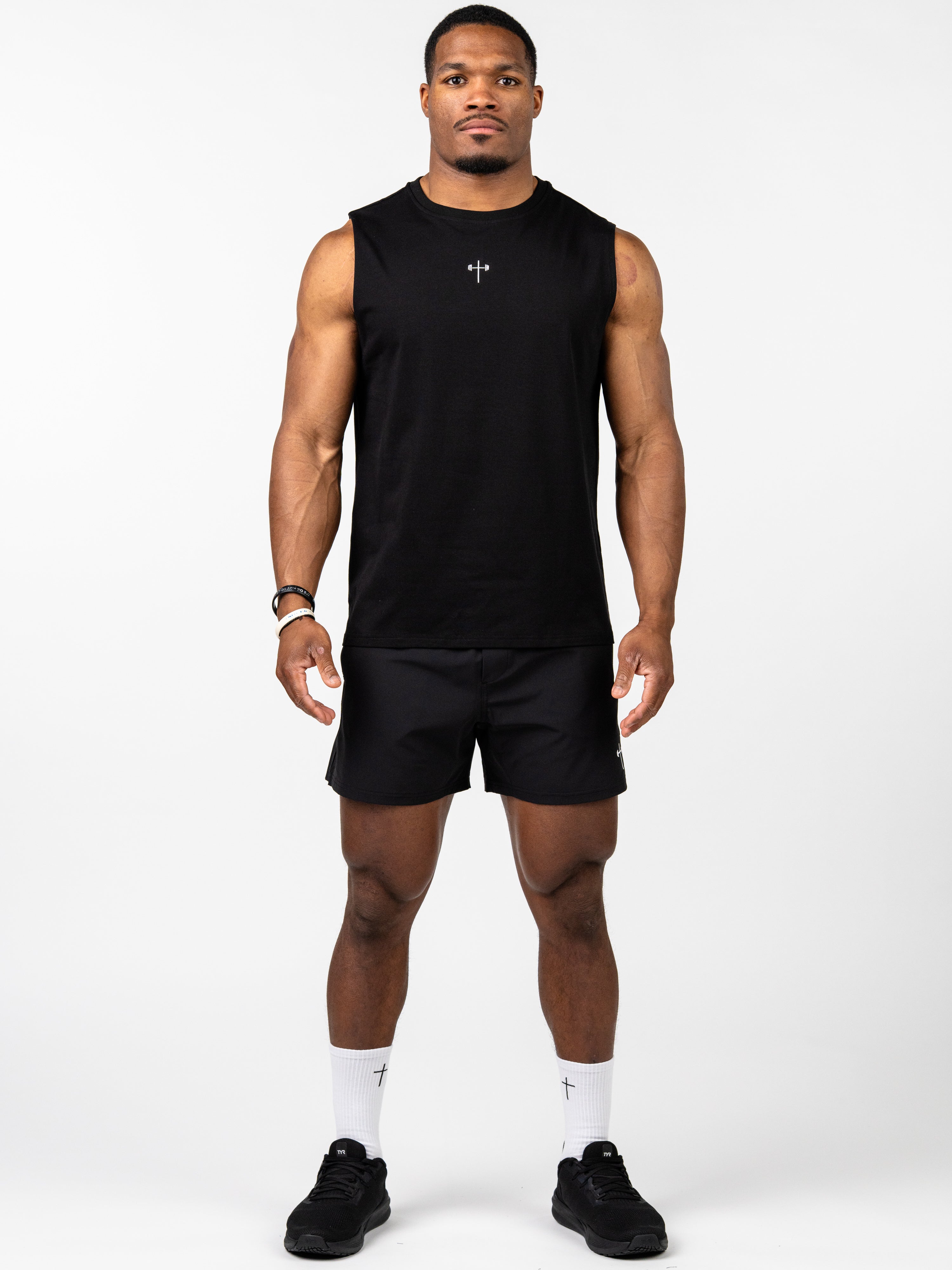 Man wearing a black Vital Cross Sleeveless Tank with a small cross and barbell symbol on the chest, paired with black athletic shorts, white socks featuring a cross, and black sneakers.