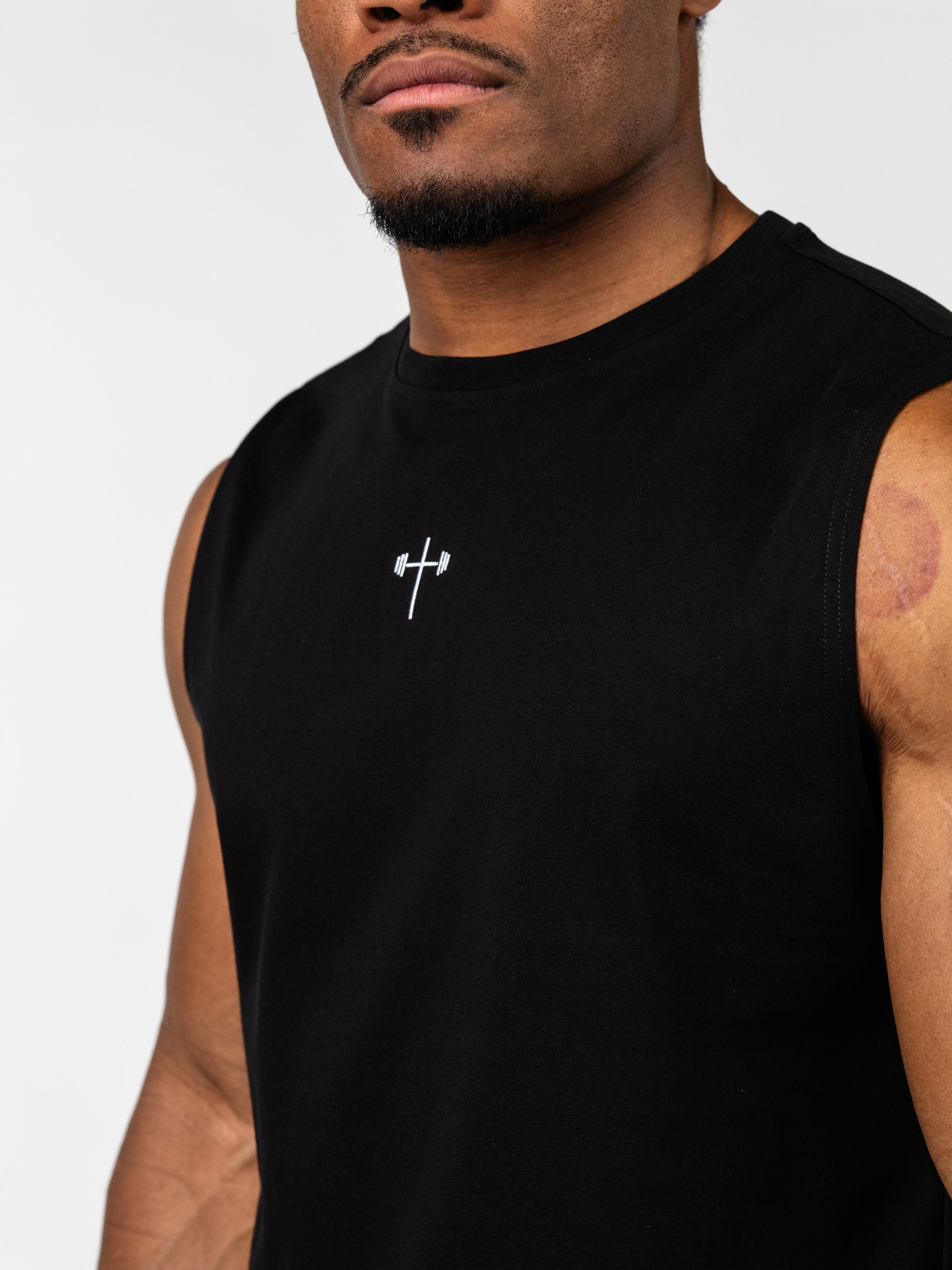Close-up of a man wearing a black Vital Cross Sleeveless Tank featuring a subtle white Christian cross with a barbell design, representing faith-based fitness apparel.