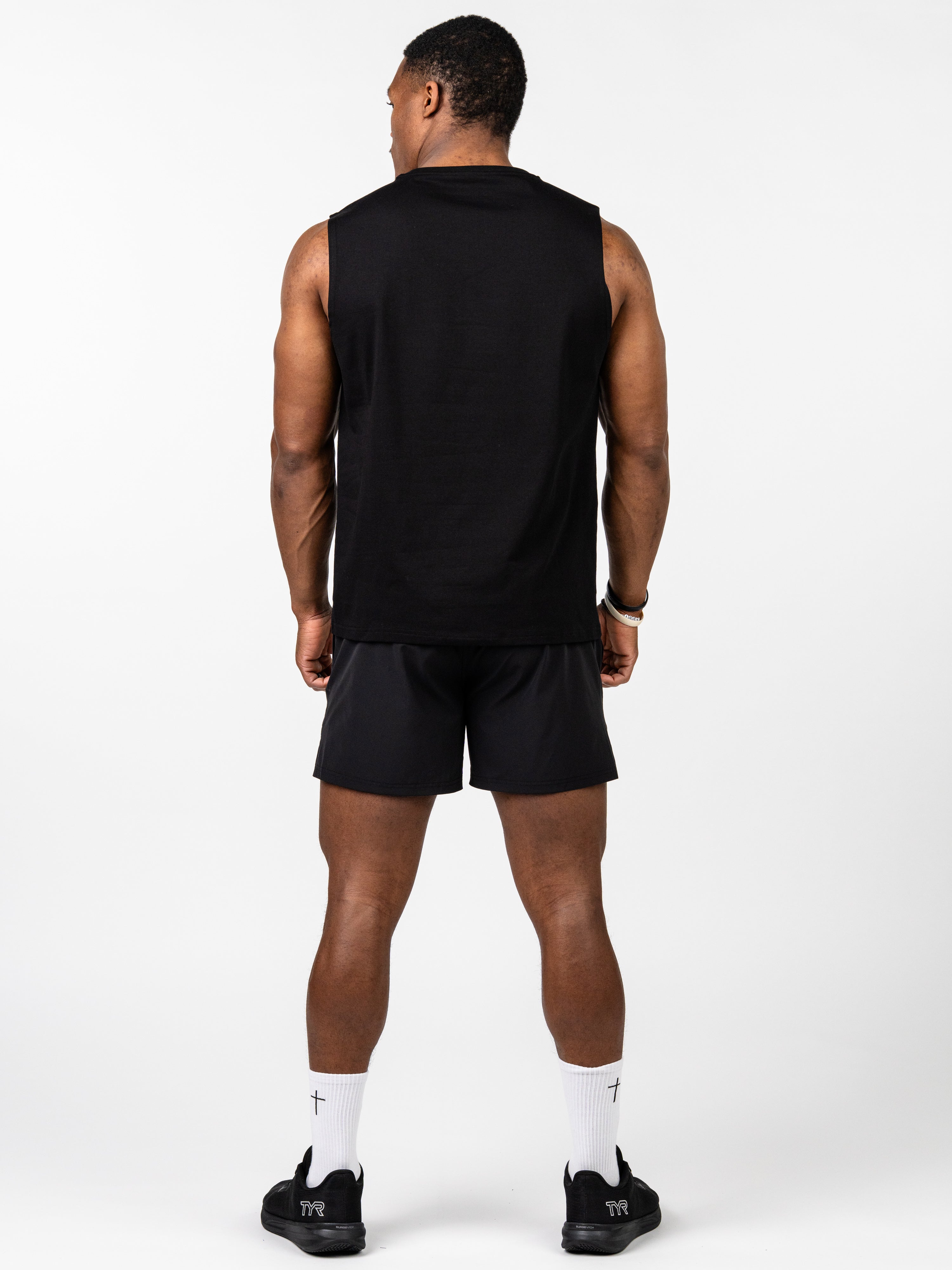 Male athlete wearing a black Vital Cross Sleeveless Tank showing the back view, paired with black shorts, white socks with cross symbols, and black athletic shoes, standing against a white background.