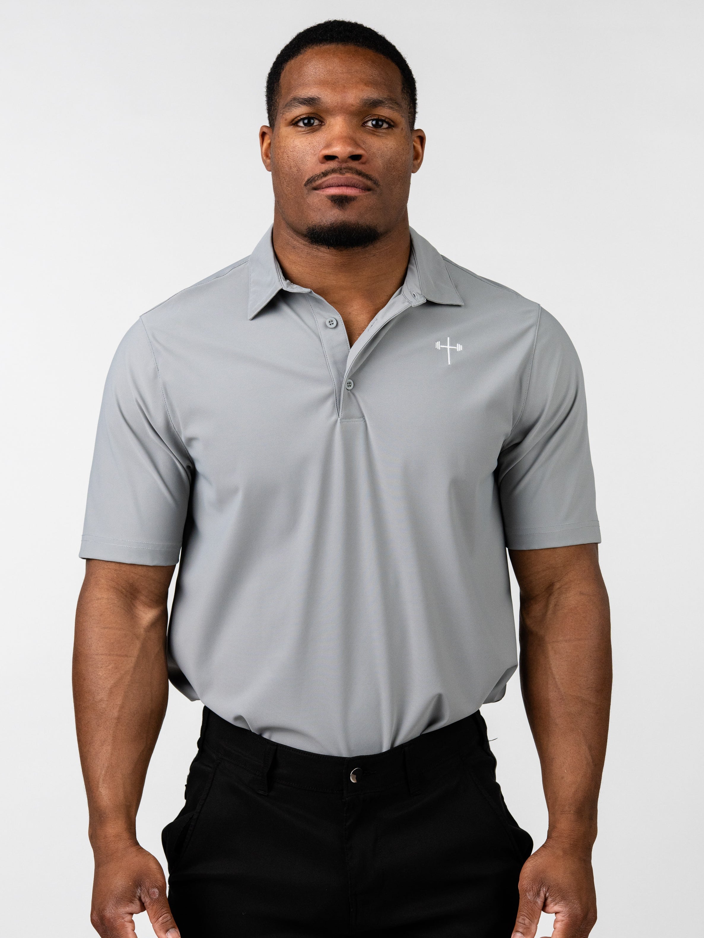 Man wearing a light gray Core Athletic Polo with a subtle Christian cross and barbell design on the left chest, paired with black pants, standing against a plain white background.