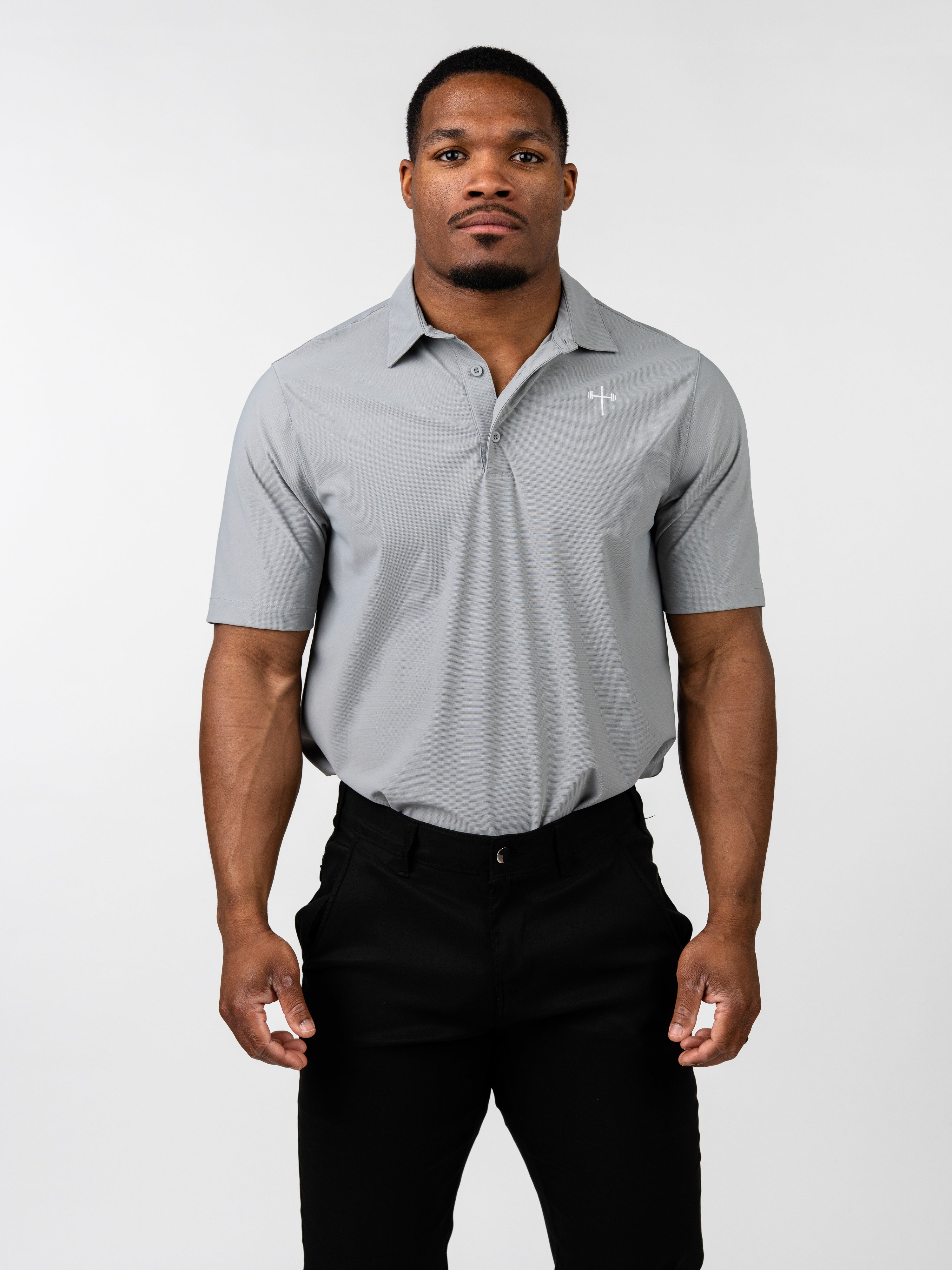 Man wearing a light gray Core Athletic Polo with a subtle Christian cross and barbell design on the left chest, paired with black pants, standing against a plain white background.