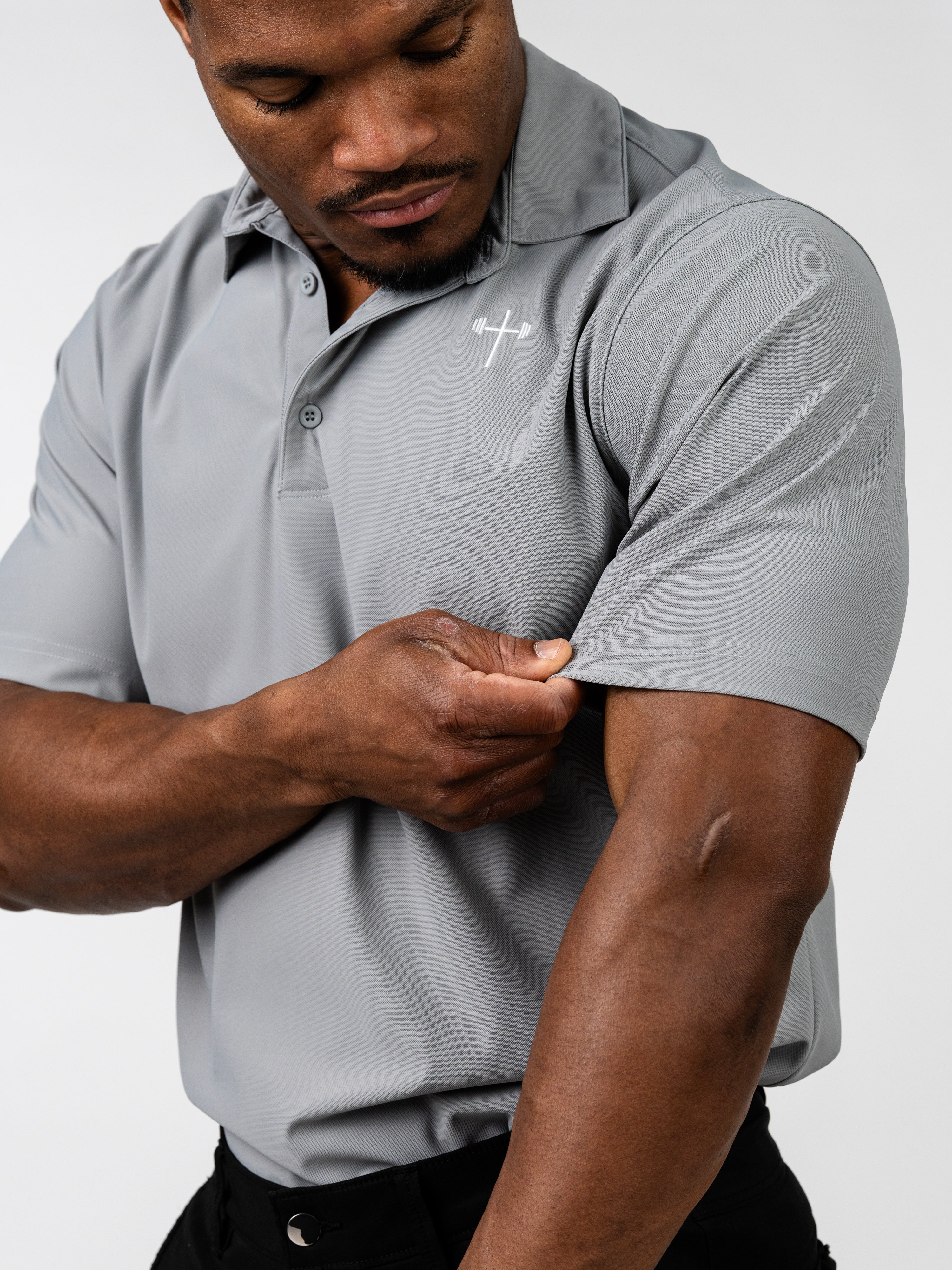 Man wearing a gray Core Athletic Polo with a subtle white Christian cross and barbell logo on the chest, demonstrating the fabric’s stretch and fit on the sleeve.