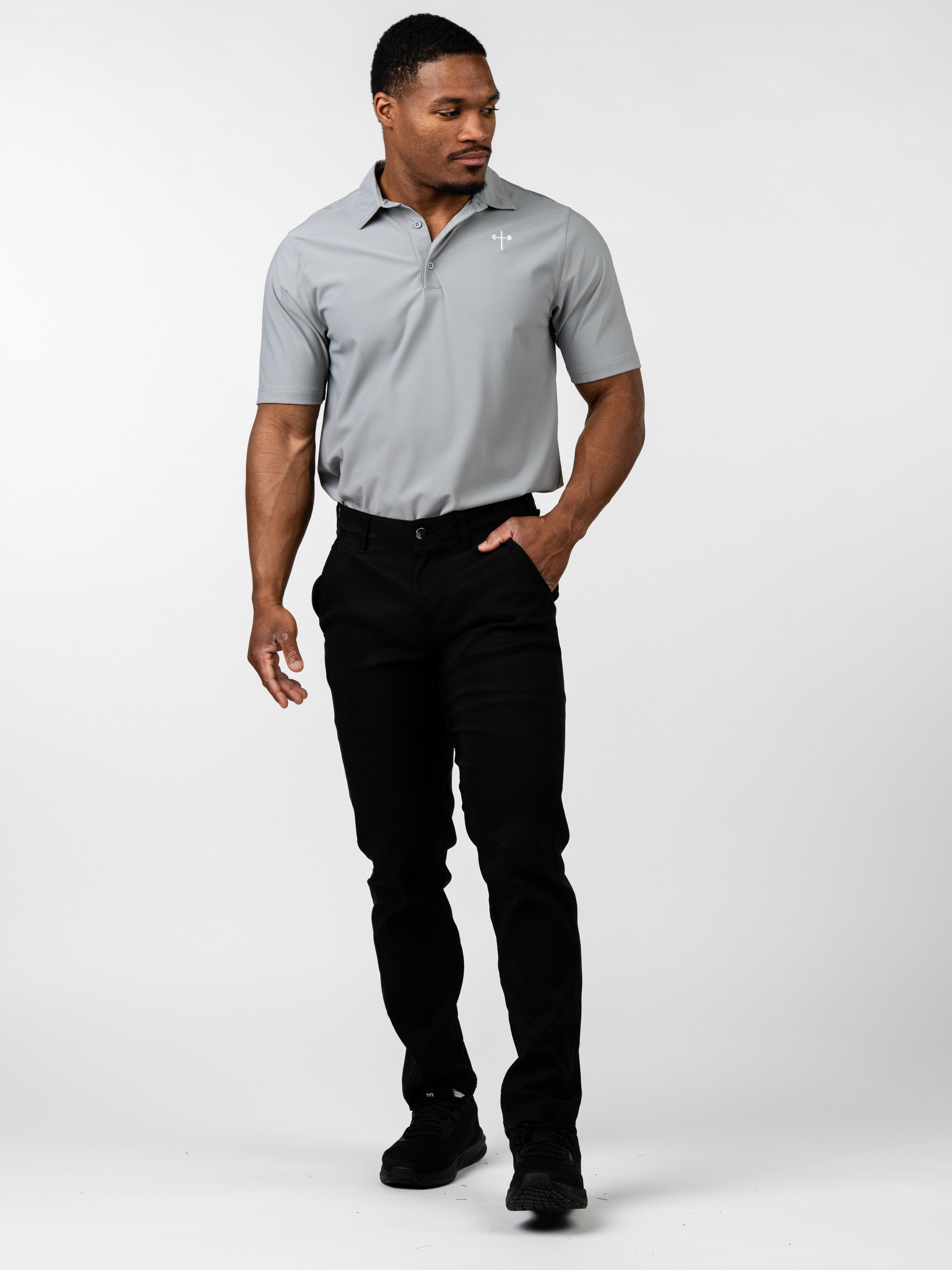 Man wearing a gray Core Athletic Polo with a subtle Christian cross logo on the chest, paired with black pants and black shoes, walking confidently against a white background.