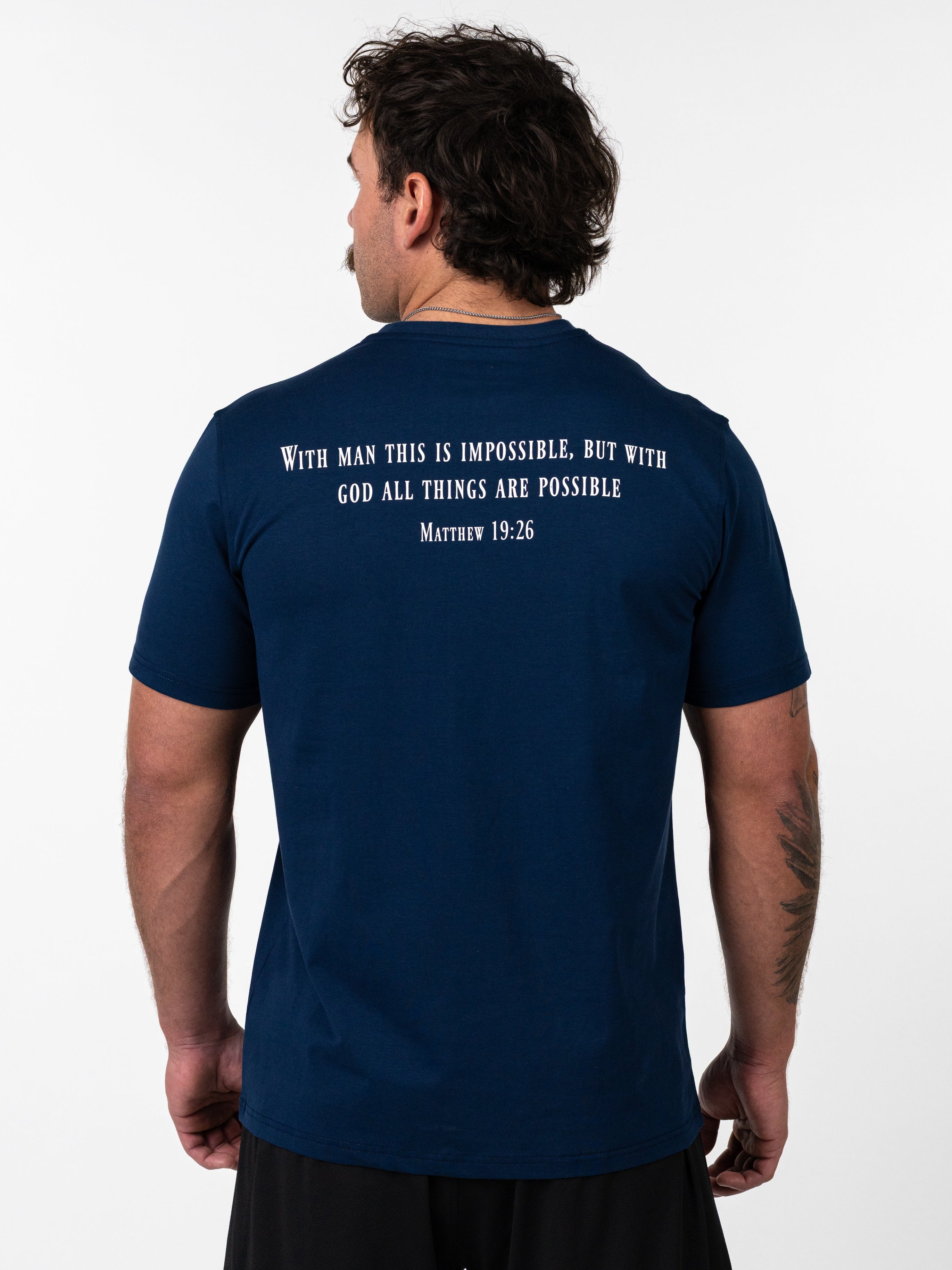 Man wearing a navy blue Sovereign Performance Tee with a Bible verse from Matthew 19:26 printed on the back, standing against a white background, showcasing Christian fitness apparel.