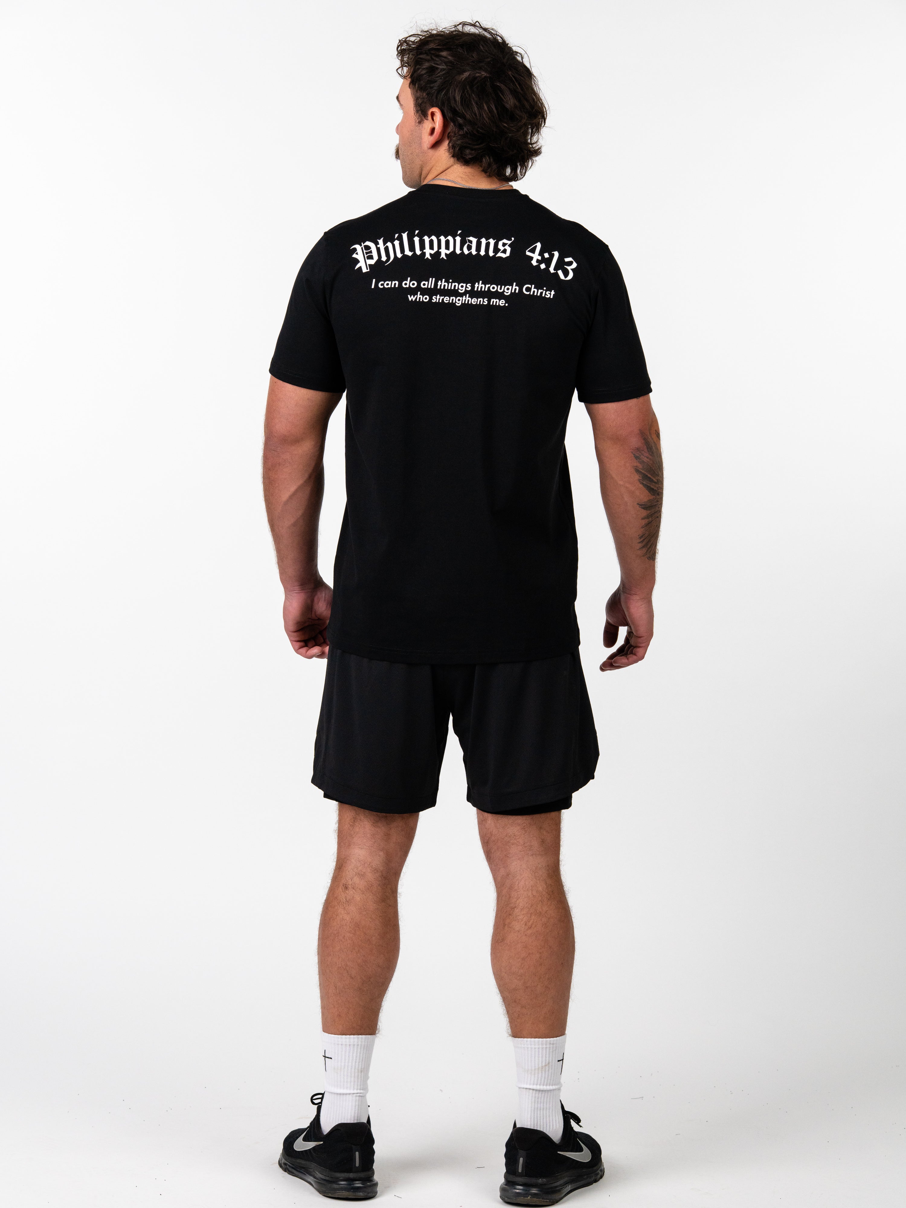 Man wearing a black Kingdom Performance Tee with Philippians 4:13 scripture printed on the back, paired with black athletic shorts, white socks, and black sneakers, shown from behind.