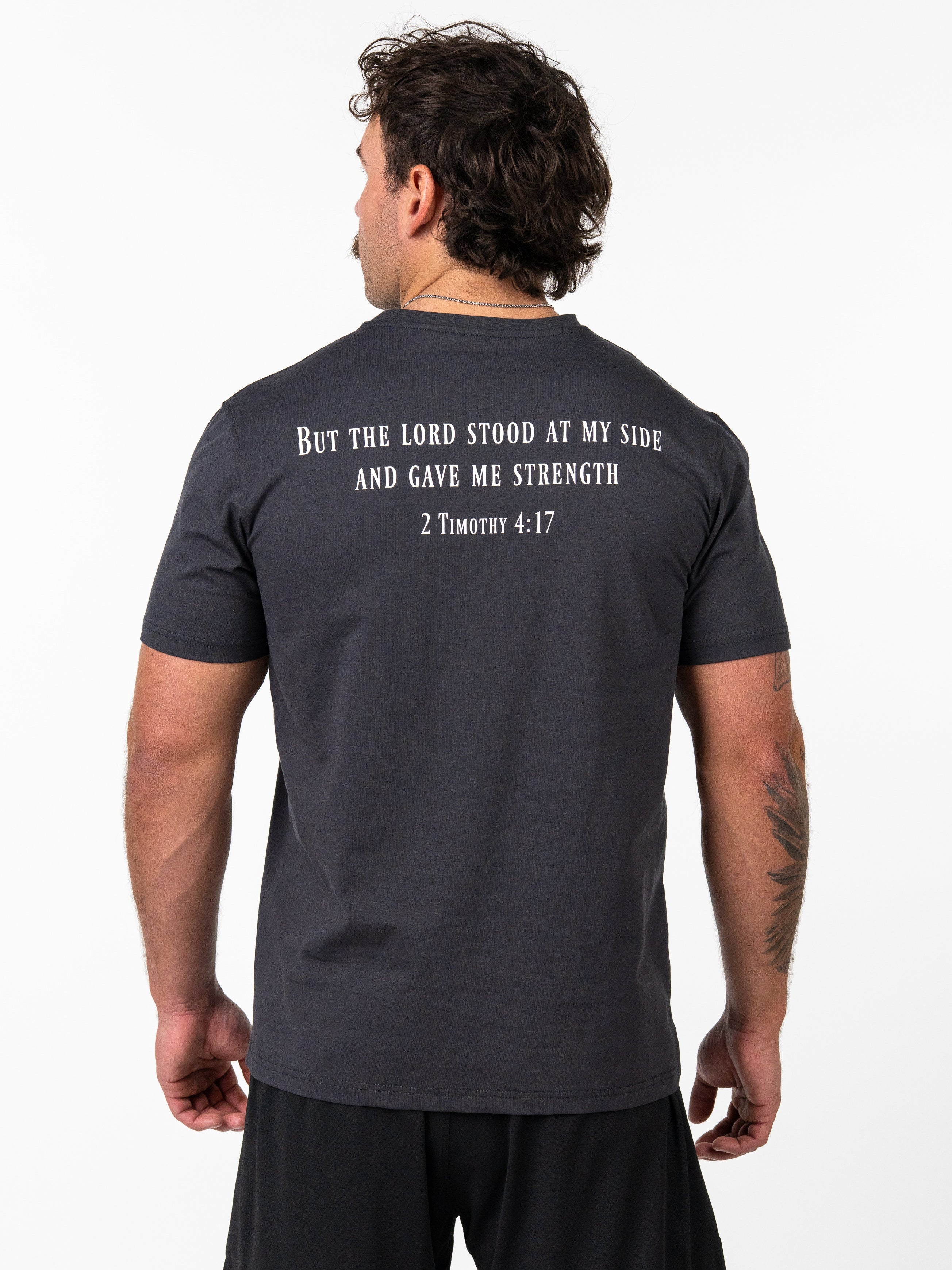 Man wearing a black Sovereign Performance Tee with a biblical quote from 2 Timothy 4:17 printed on the back, paired with black athletic shorts in a studio setting.