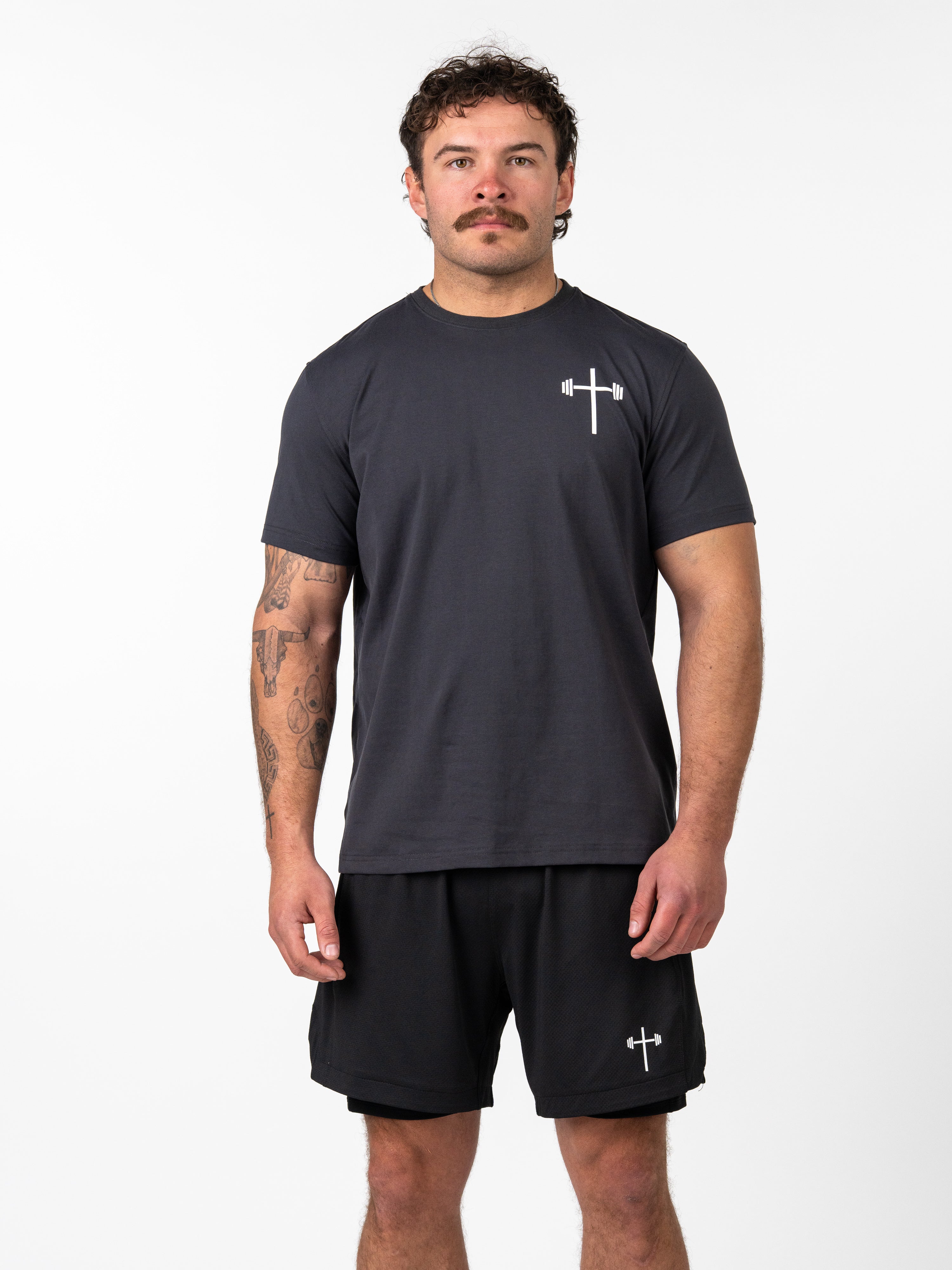 Man wearing a black Kingdom Performance Tee featuring a small white cross with a barbell design on the left chest, paired with black shorts, standing against a white background.