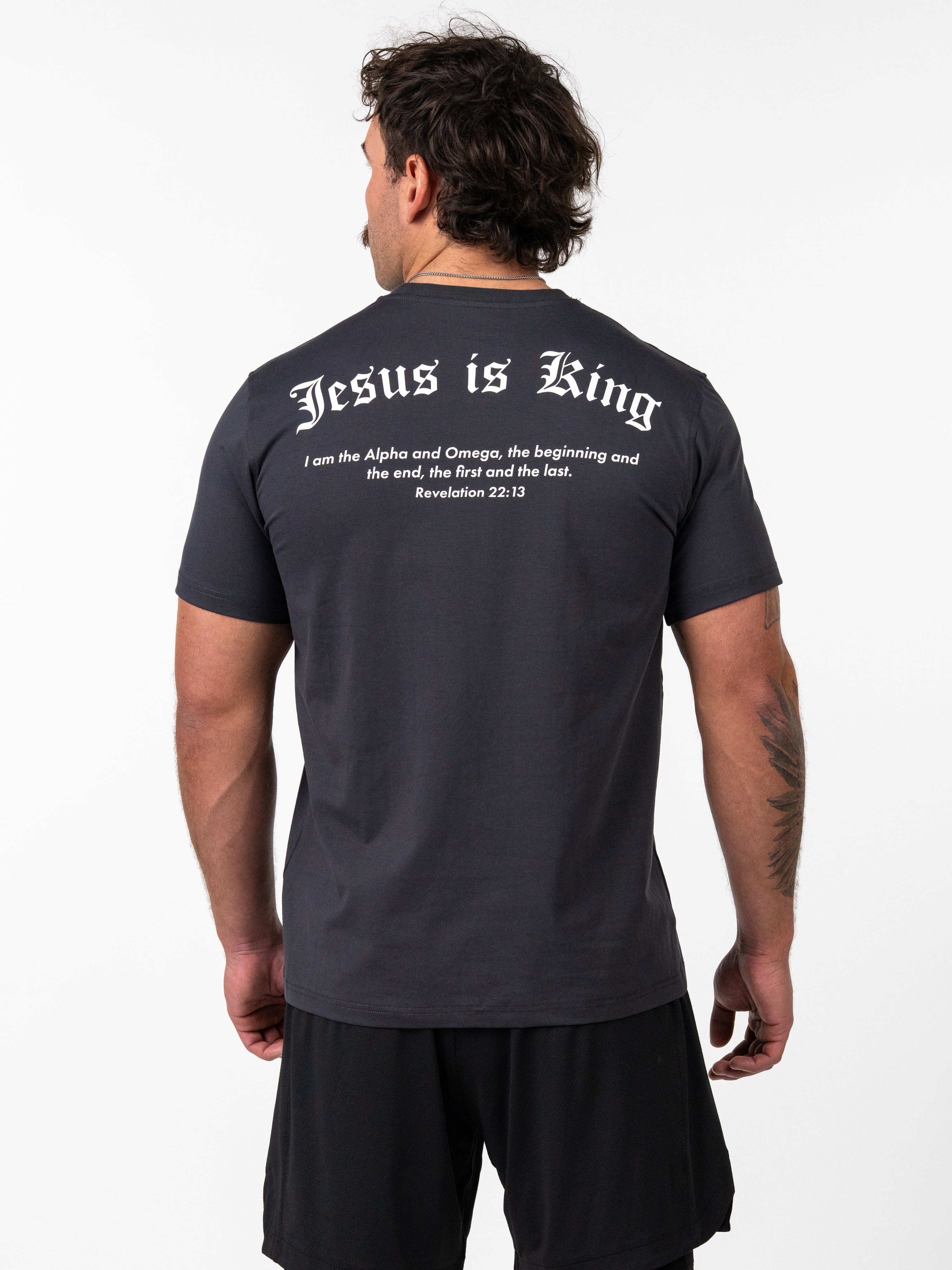 Man wearing a black Kingdom Performance Tee with the back printed text "Jesus is King" and a Bible verse from Revelation 22:13, shown in a faith-based fitness apparel style.