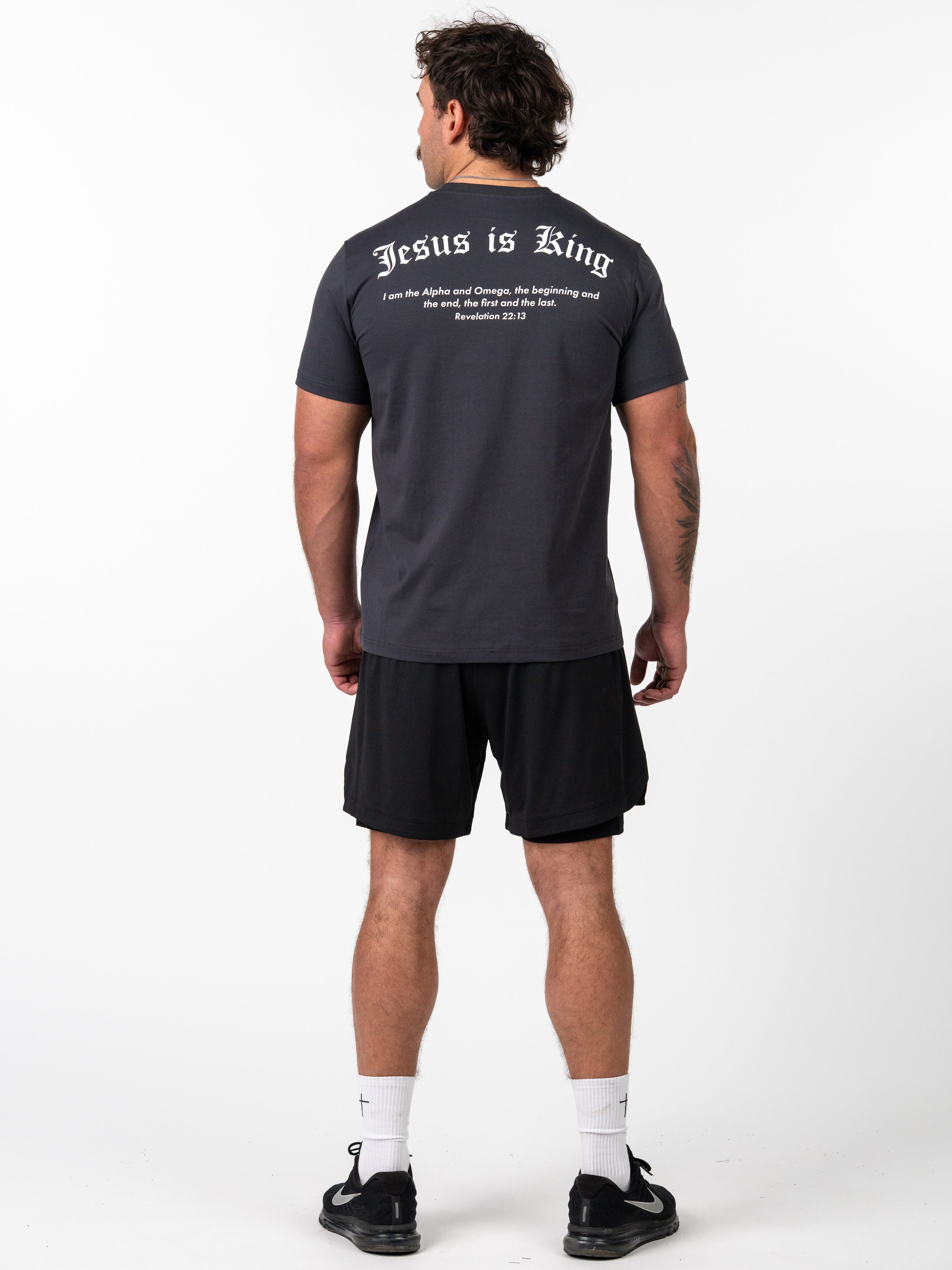 Man wearing a black Kingdom Performance Tee featuring "Jesus is King" text and a Bible verse on the back, paired with black athletic shorts, white socks, and black sneakers.