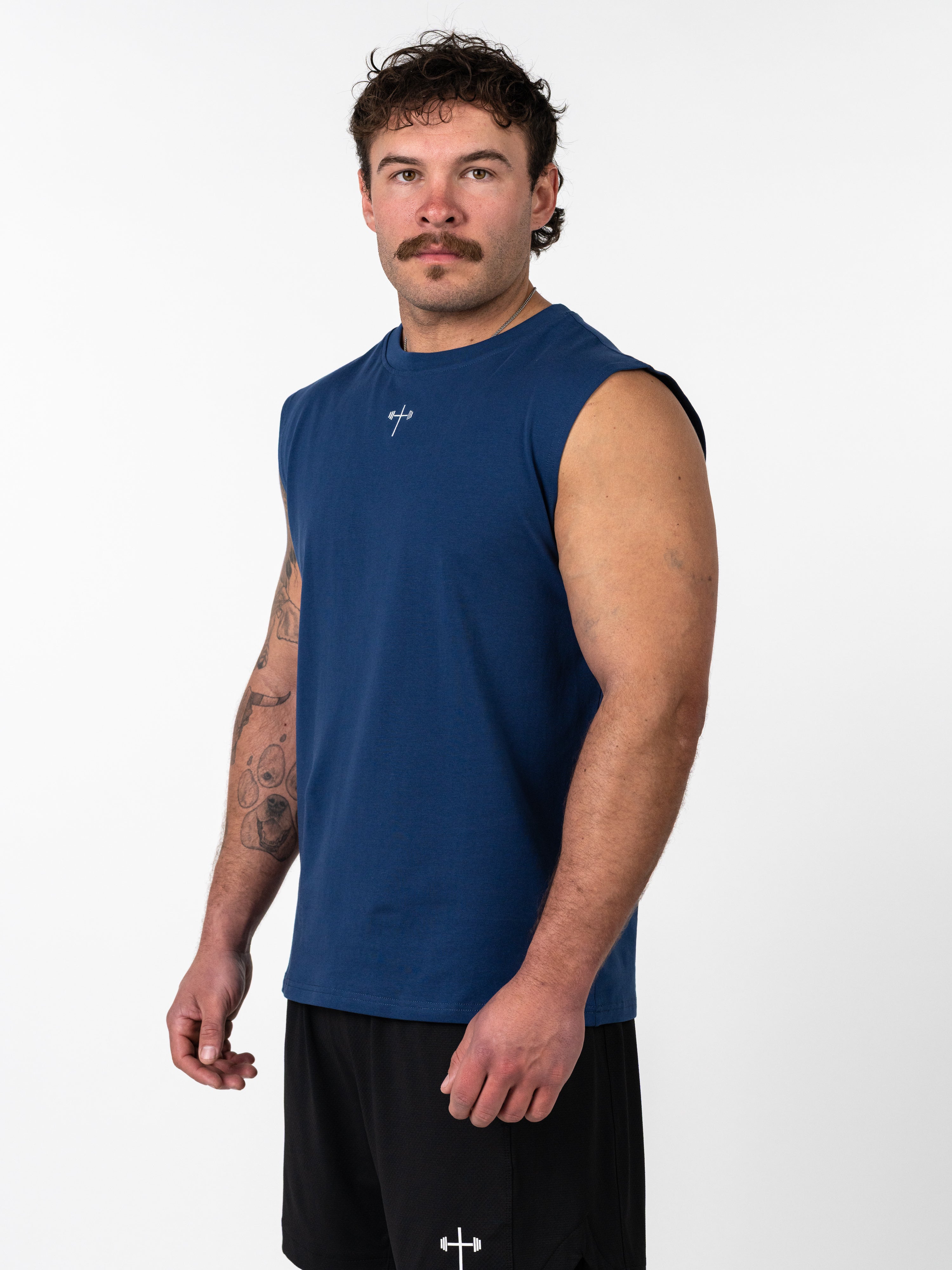 Man wearing a navy blue Scripture Premium Sleeveless Tank with a small cross and barbell design on the chest, paired with black gym shorts, standing against a white background.