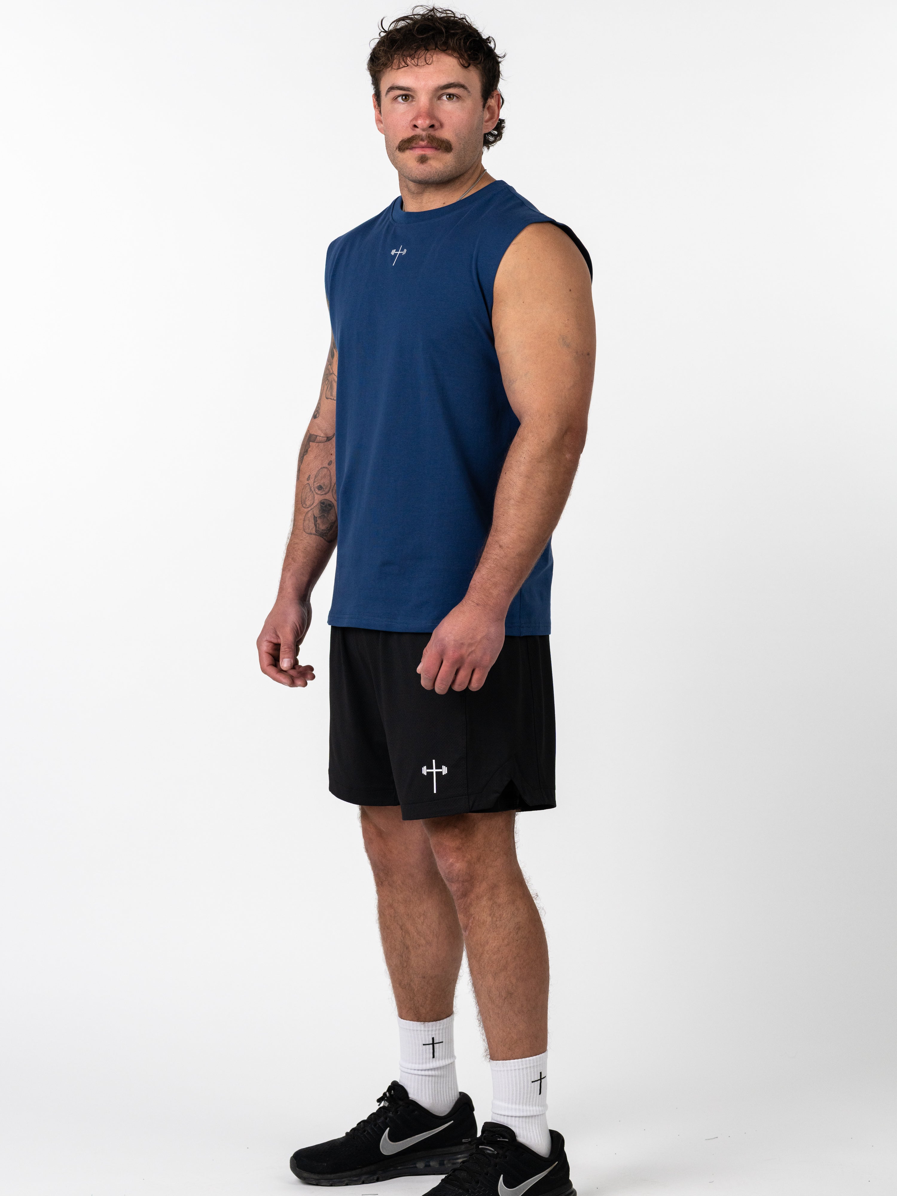 Man wearing a navy blue Scripture Premium Sleeveless Tank paired with black shorts featuring a small cross and barbell graphic, standing against a white background.