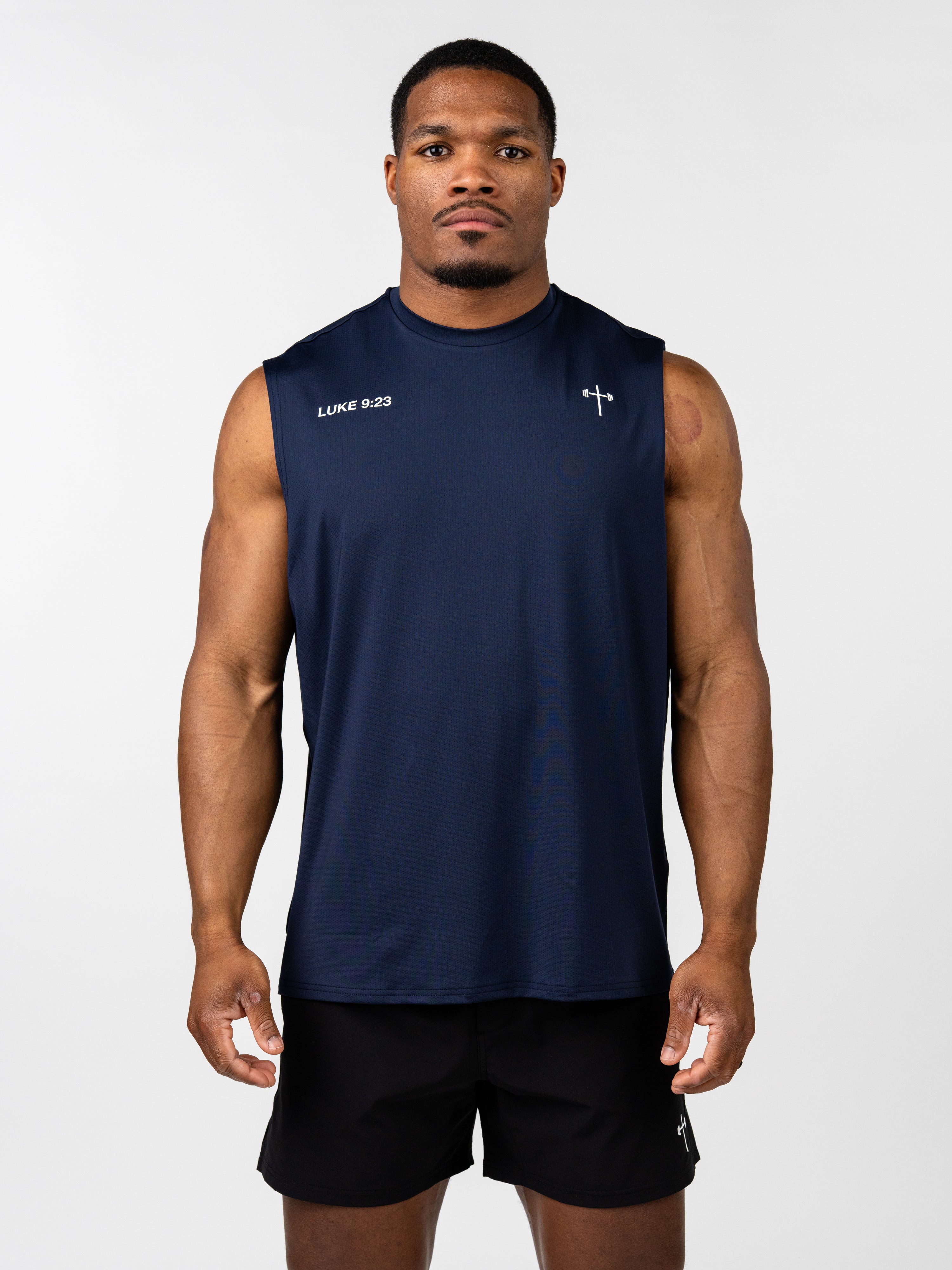 Man wearing navy blue Carry Your Cross Premium Oversized Tank with sleeveless design and small cross and "Luke 9:23" text on chest, paired with black shorts.