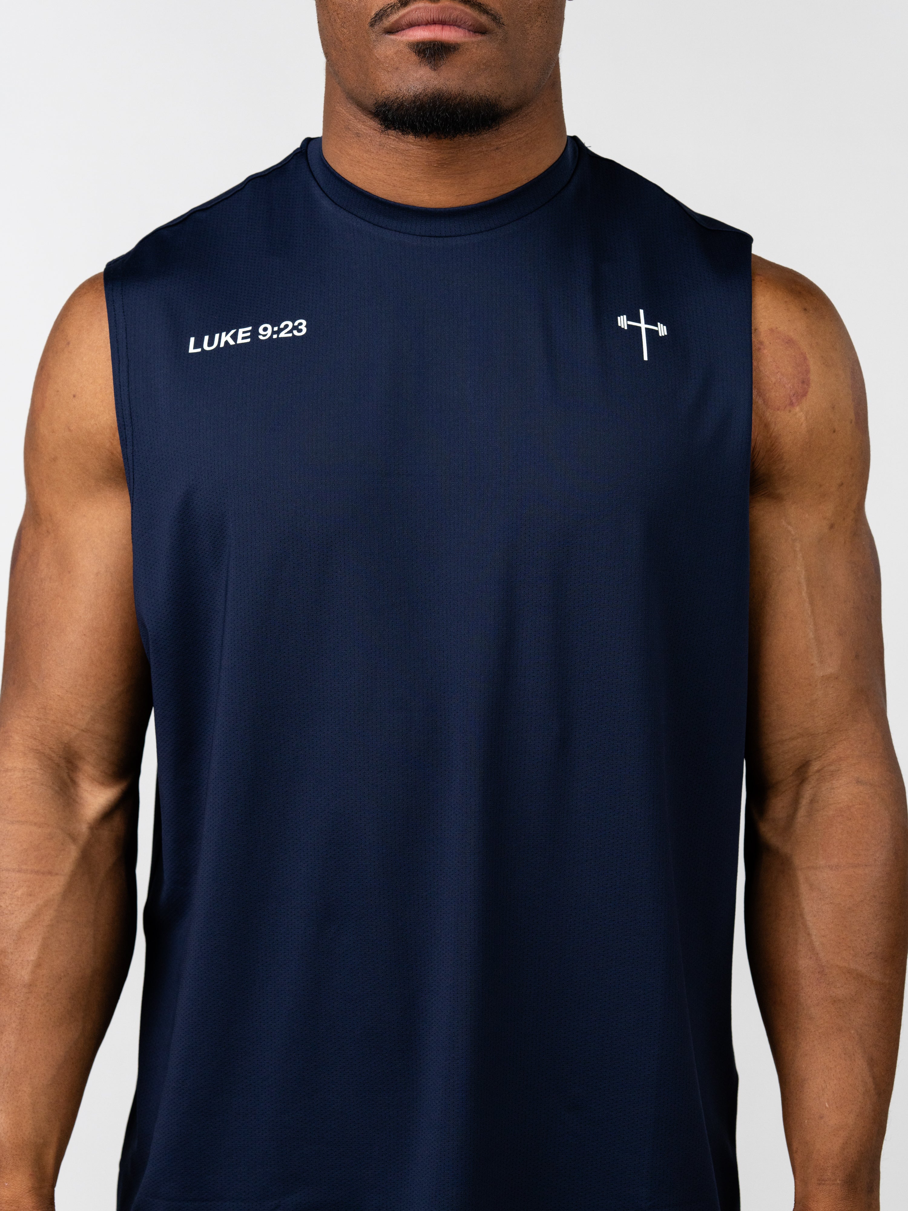 Man wearing a navy blue sleeveless athletic tank top featuring "Carry Your Cross Premium Oversized Tank" with white "LUKE 9:23" text and a small cross with barbell graphic on the chest, designed for Christian fitness apparel.