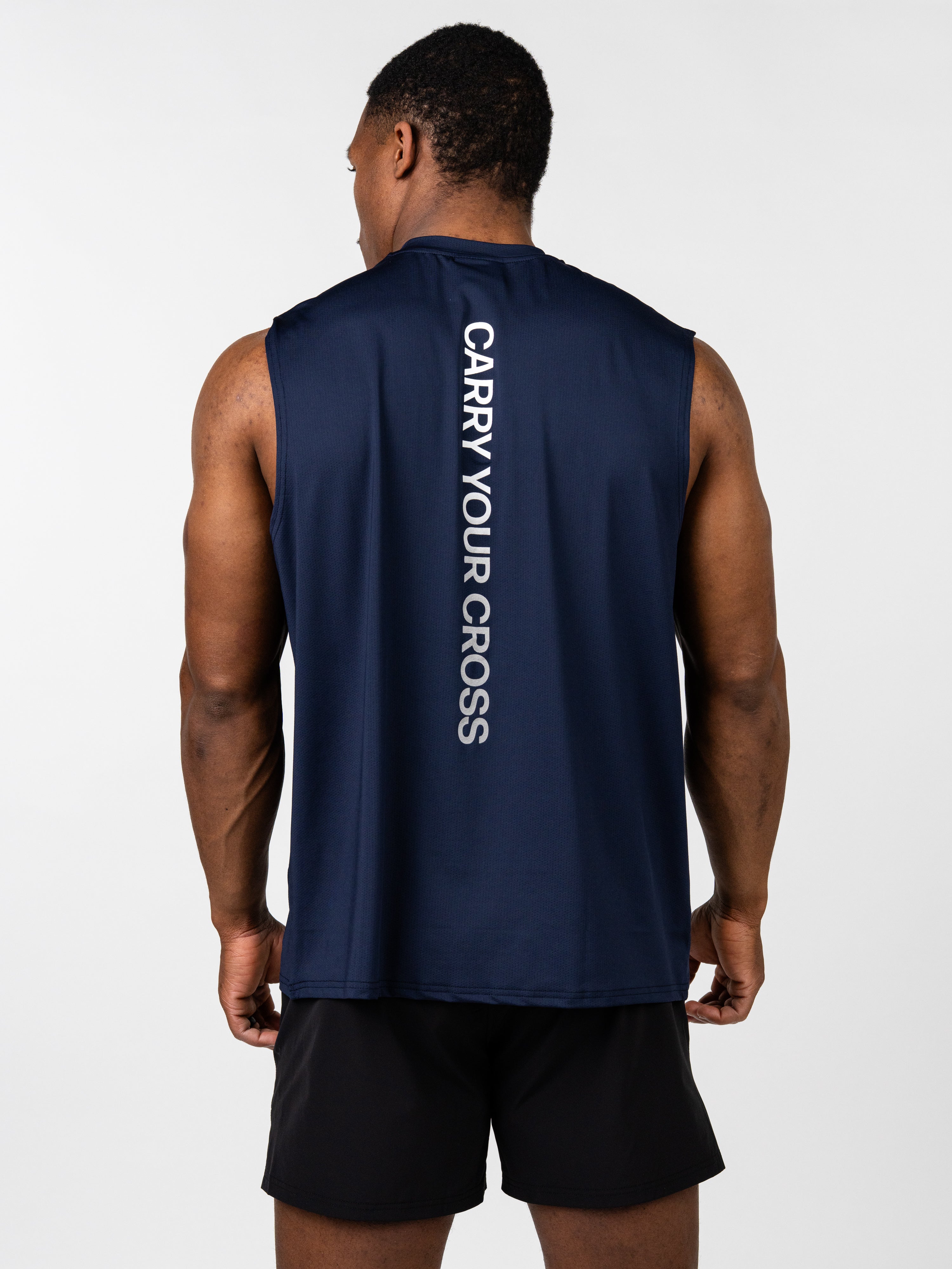 Man wearing a navy blue Carry Your Cross Premium Oversized Tank with white vertical text on the back, paired with black athletic shorts, showcasing a faith-based fitness apparel style.