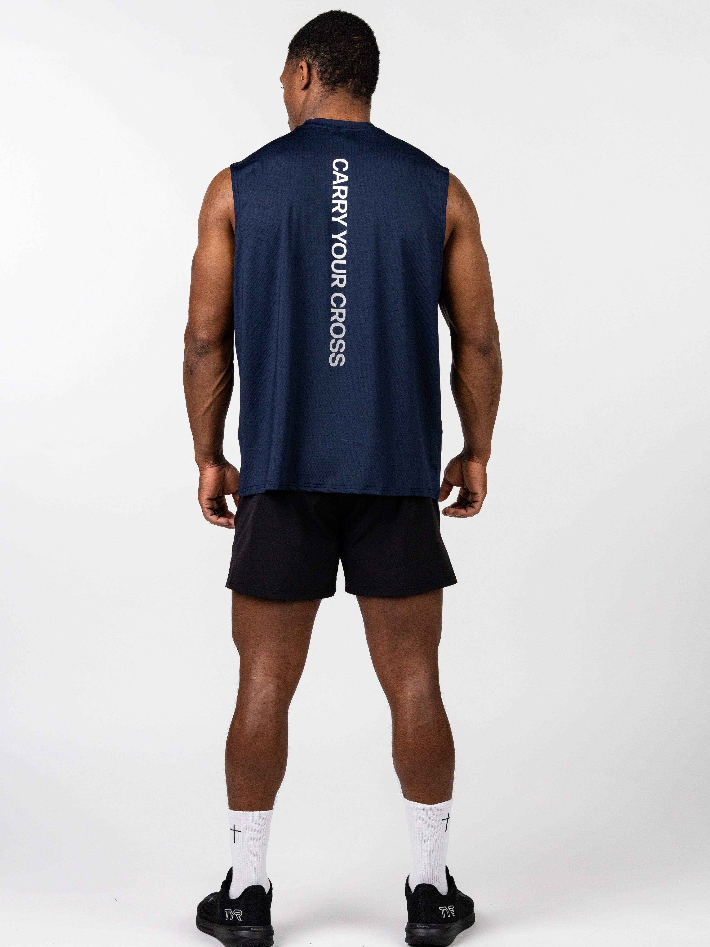 Man wearing a navy blue Carry Your Cross Premium Oversized Tank with vertical white lettering on the back, paired with black shorts, white socks with crosses, and black athletic shoes.