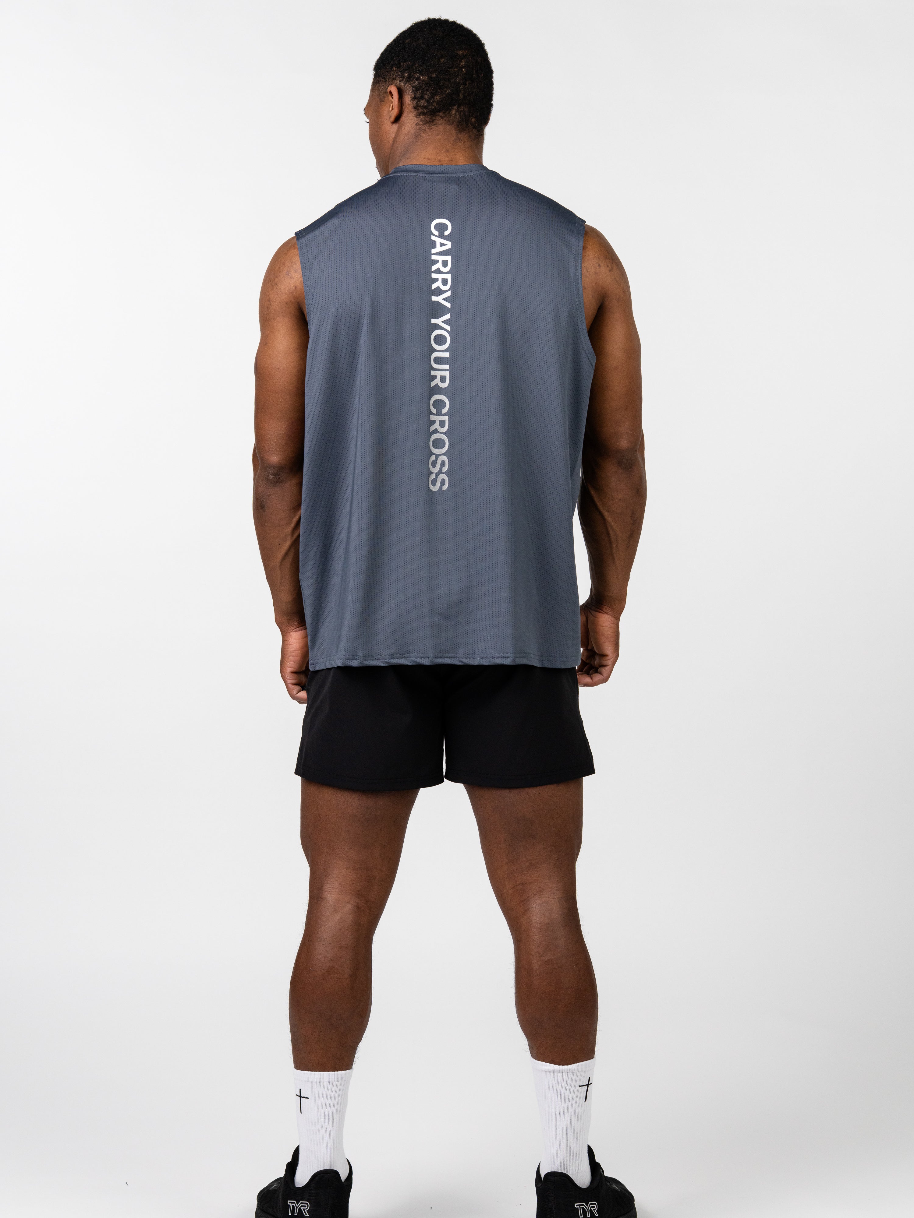 Man wearing a gray sleeveless athletic tank with "CARRY YOUR CROSS" printed vertically on the back, paired with black shorts and white socks, shown from behind wearing the Carry Your Cross Premium Oversized Tank.