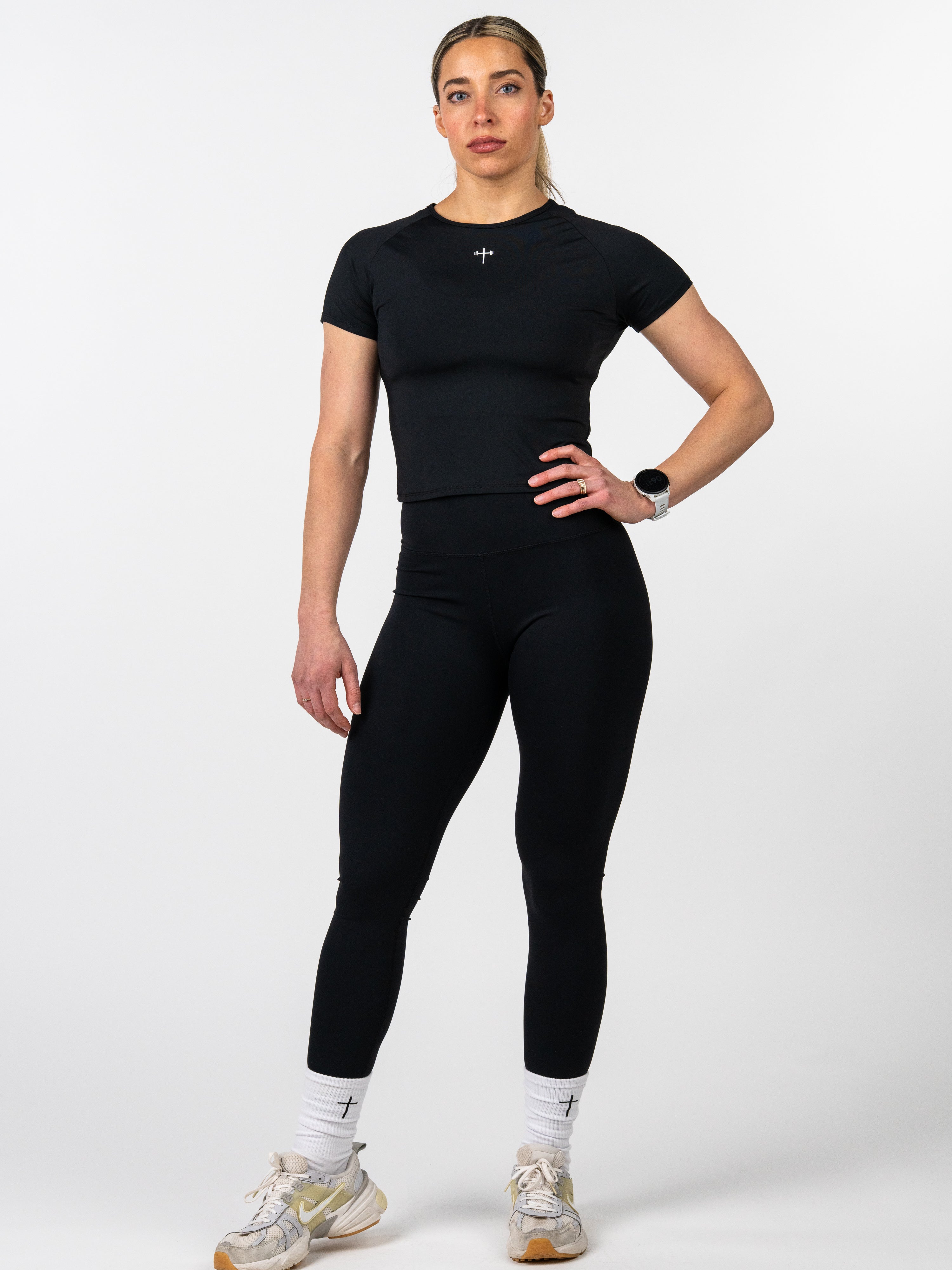 Woman wearing black Women's Combat Tee with a small cross emblem on the chest, paired with black leggings, white socks featuring cross symbols, and athletic shoes.