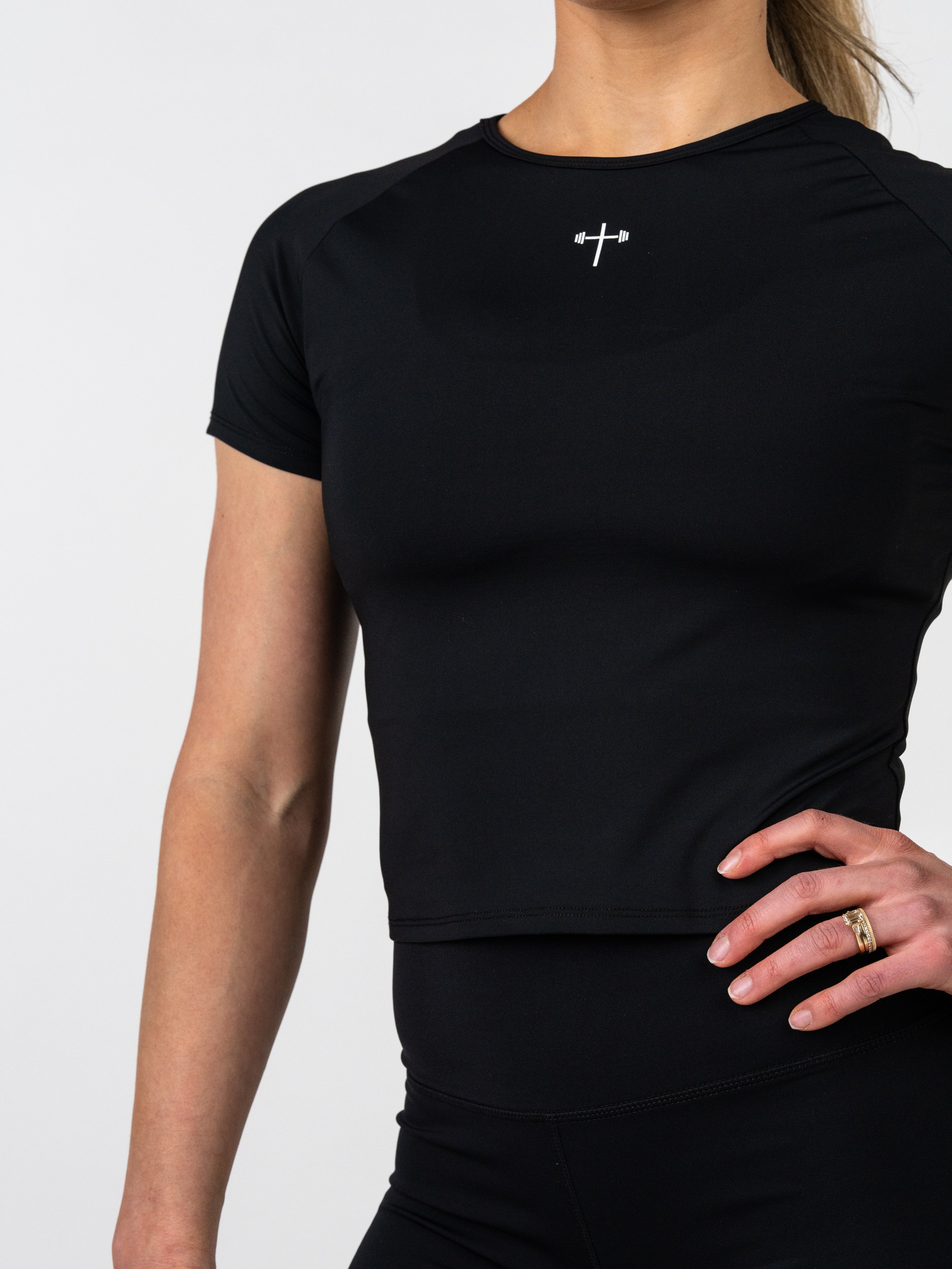 Close-up of a woman wearing a black Women's Combat Tee featuring a small white cross with a barbell design on the chest, designed for performance and movement.