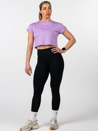Woman wearing a lavender Cross Essential Crop Tee with a small Christian cross and barbell design on the chest, paired with black leggings, white socks with crosses, and athletic shoes, modeling Christian fitness apparel.