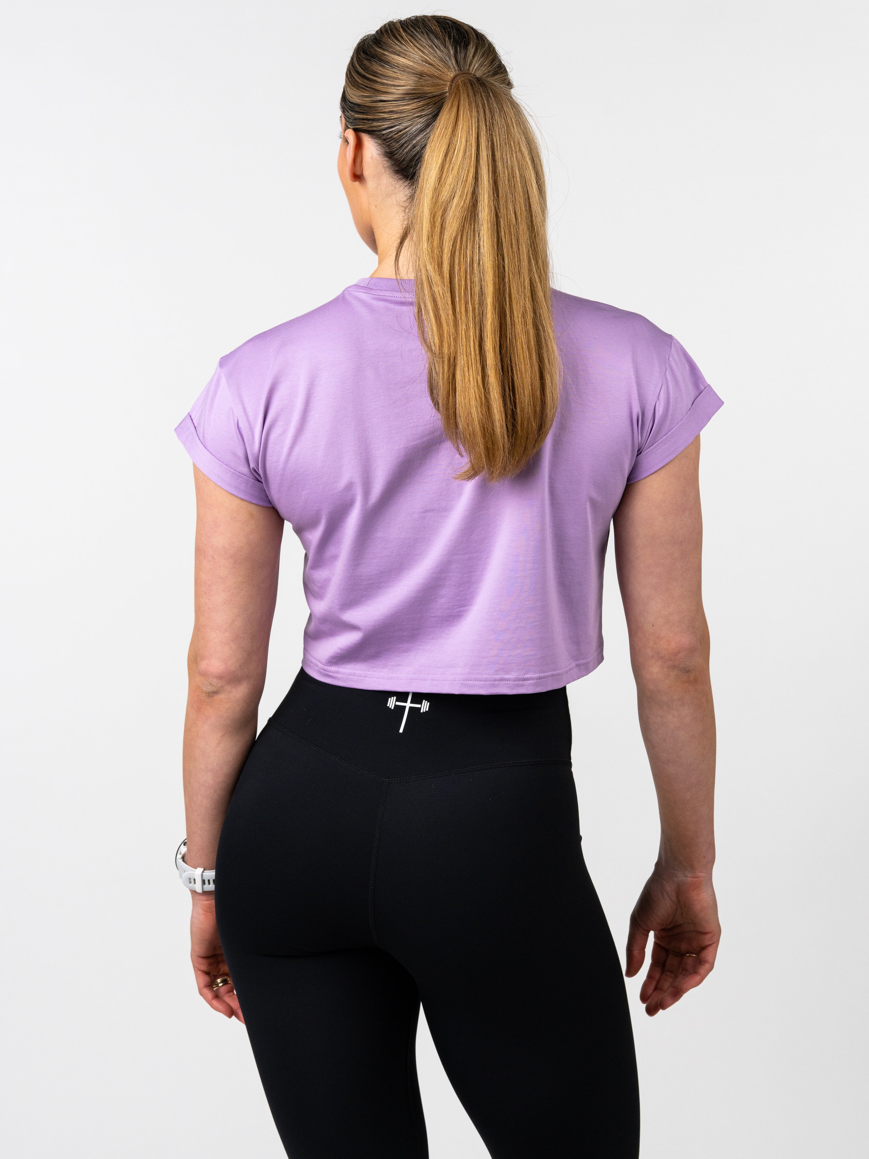 Woman wearing a lavender Cross Essential Crop Tee paired with black leggings that feature a small barbell cross logo on the waistband, showcasing Christian fitness apparel.