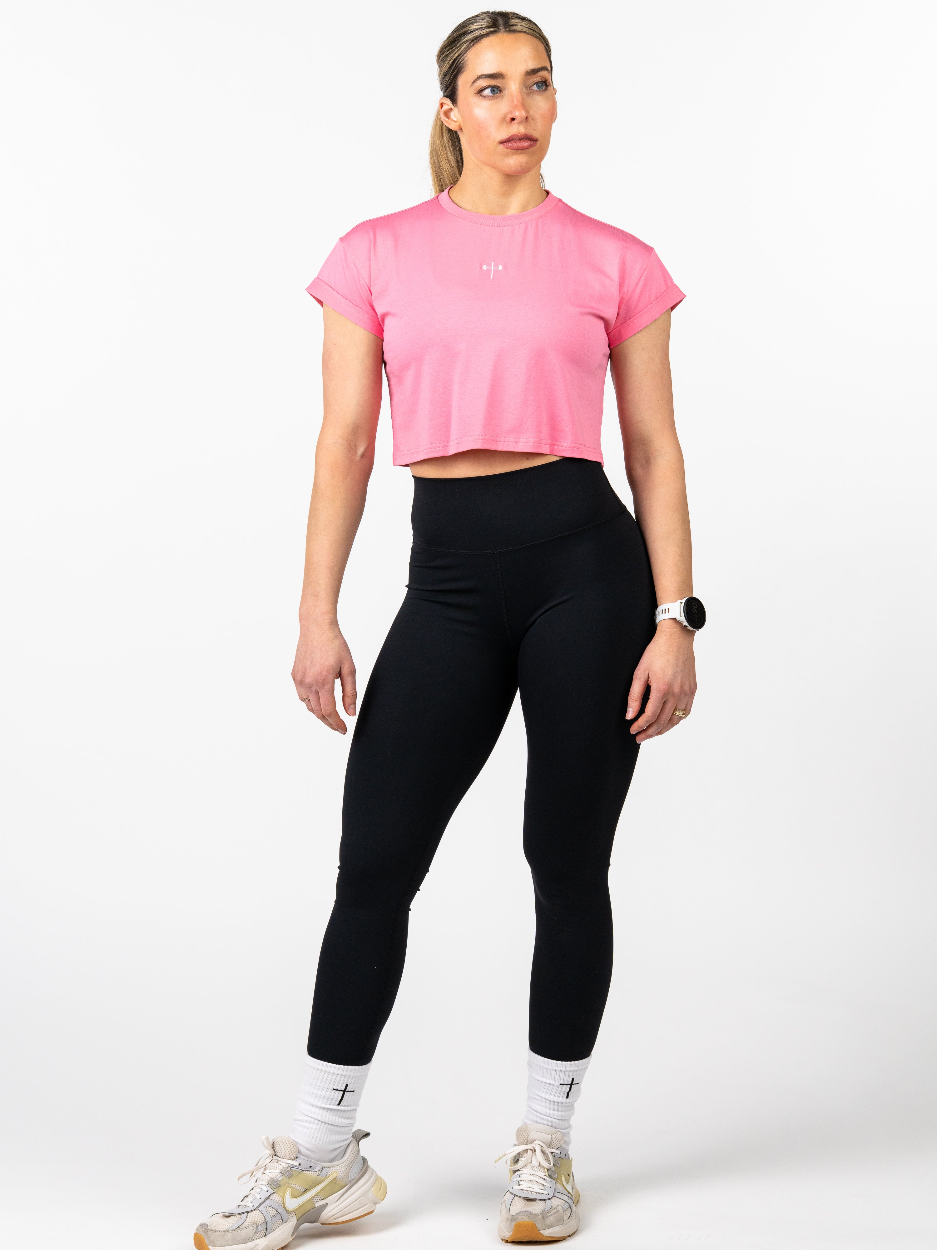 Woman wearing a pink Cross Essential Crop Tee with a small cross and barbell design on the chest, paired with black leggings and white socks featuring cross symbols, standing against a white background.
