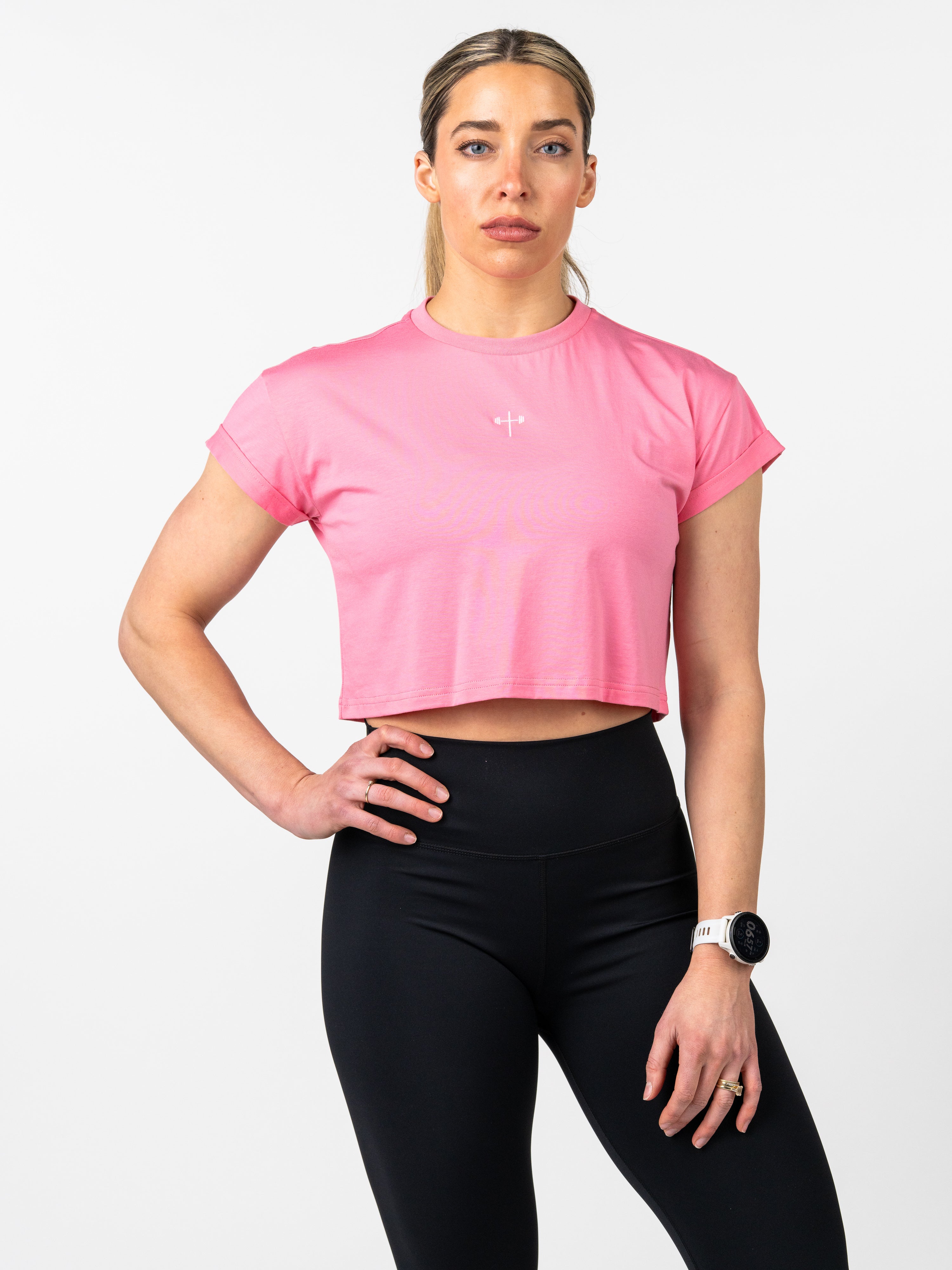 Woman wearing a pink Cross Essential Crop Tee with a subtle cross and barbell graphic, paired with black leggings and a white smartwatch, standing against a white background.